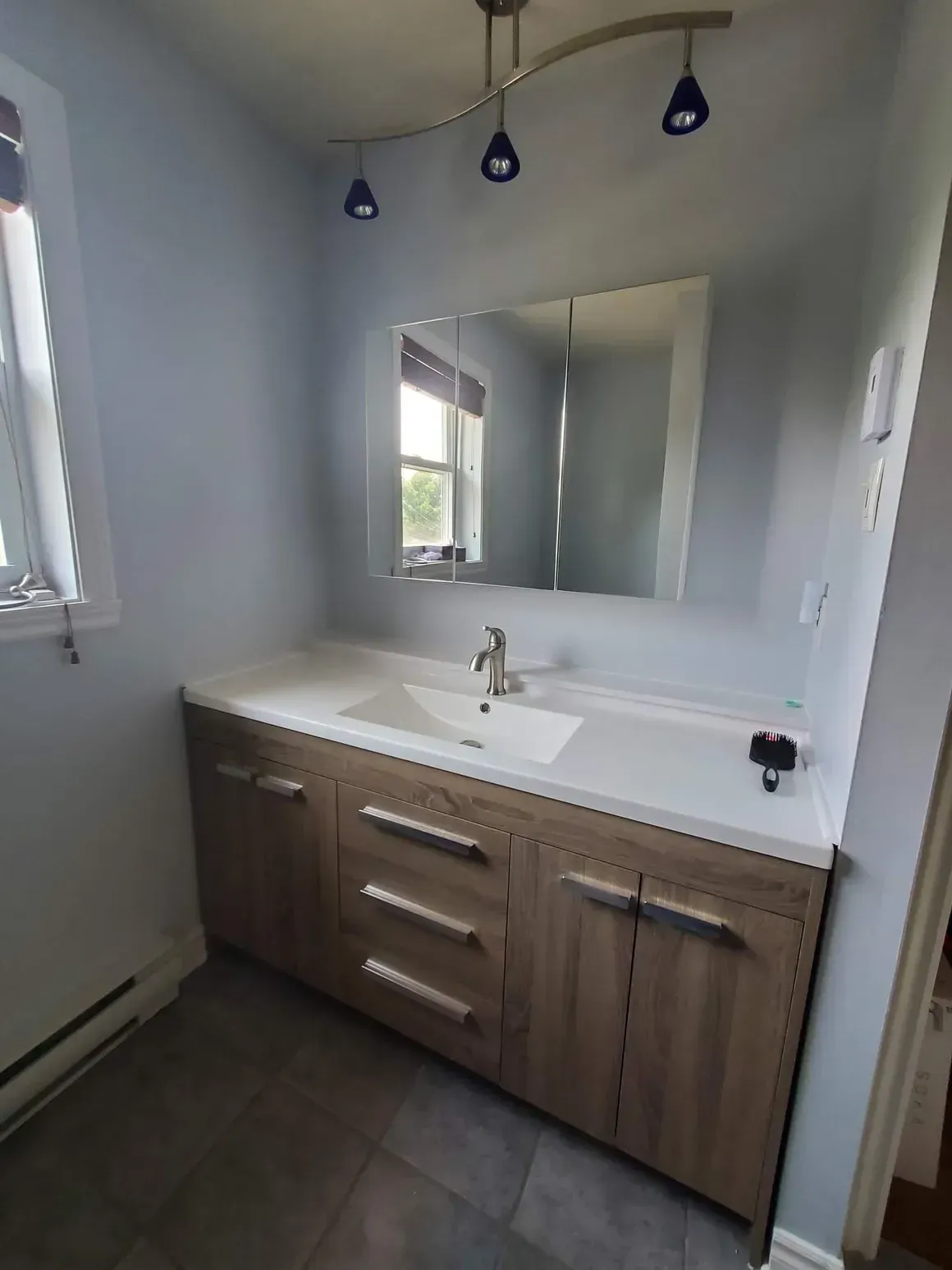 Salle de bains avec meuble-lavabo clair, comptoir blanc, grand miroir et éclairage sur rail. Murs et sol gris.