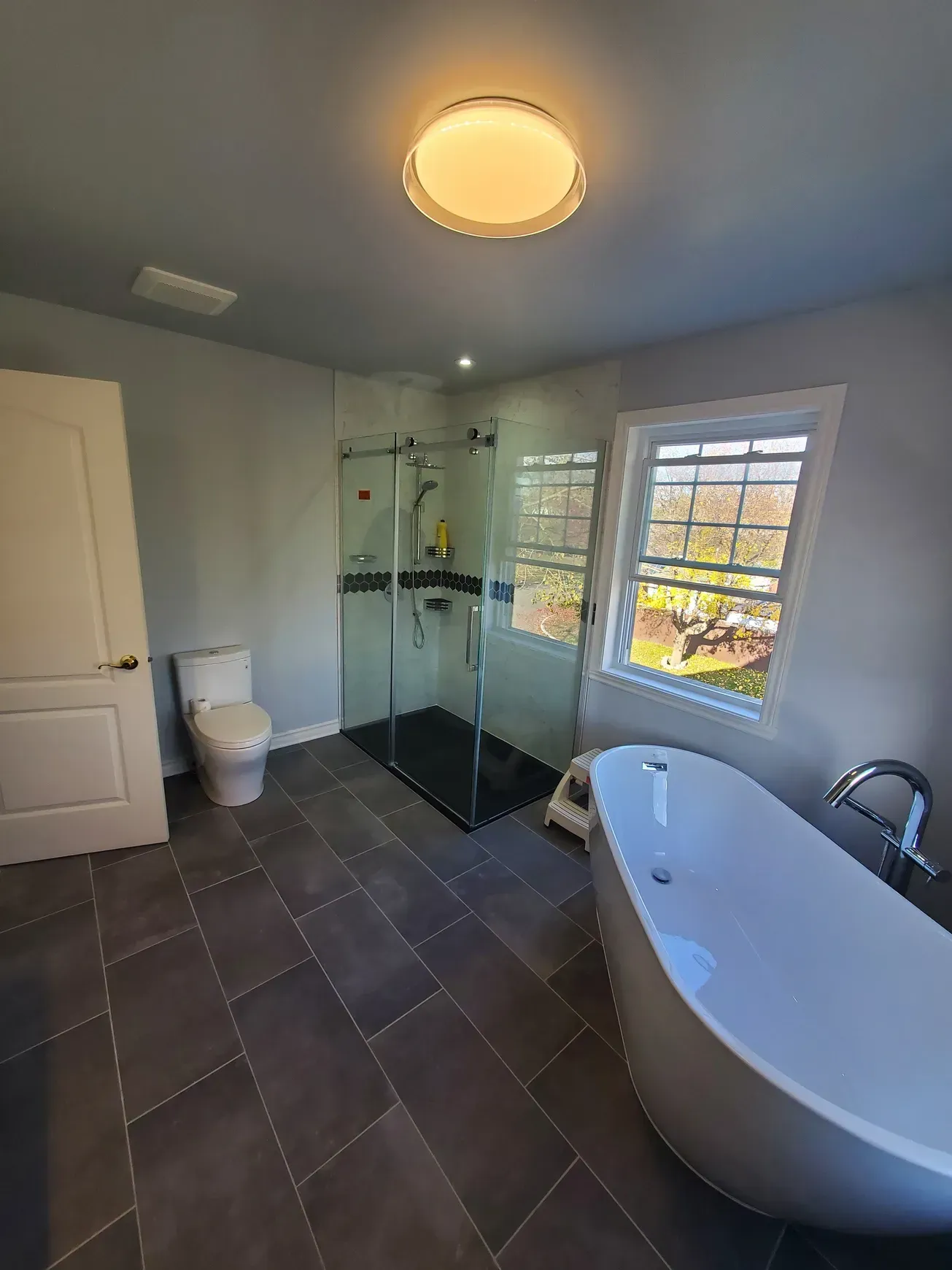 Salle de bains moderne avec douche à l'italienne, baignoire, double vasque et grande fenêtre donnant sur les arbres.