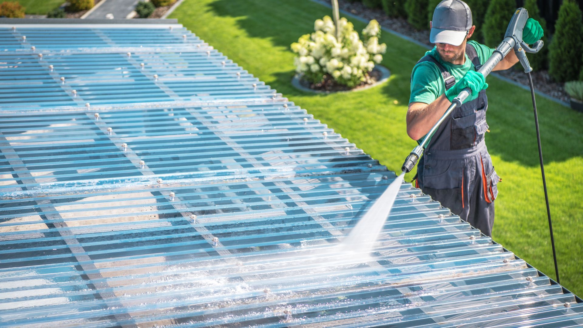 A person in work clothes uses a power washer to clean a corrugated translucent roof panel outdoors.