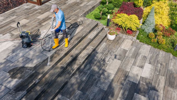 A worker in a blue shirt and yellow boots pressure-washes a stone patio next to a garden.