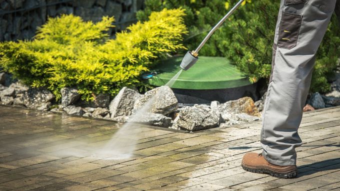 A person cleans brick pavers with a high-pressure water spray in a sunny garden setting.