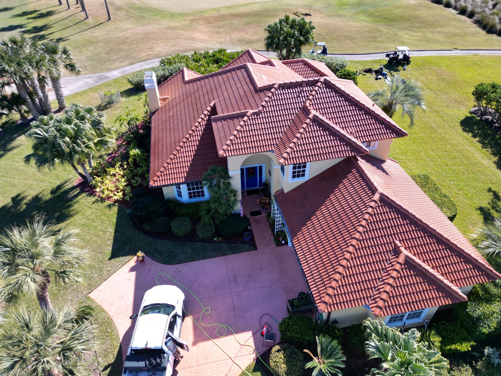Aerial view of a house with a red tile roof and a driveway, situated near a golf course with palm trees.