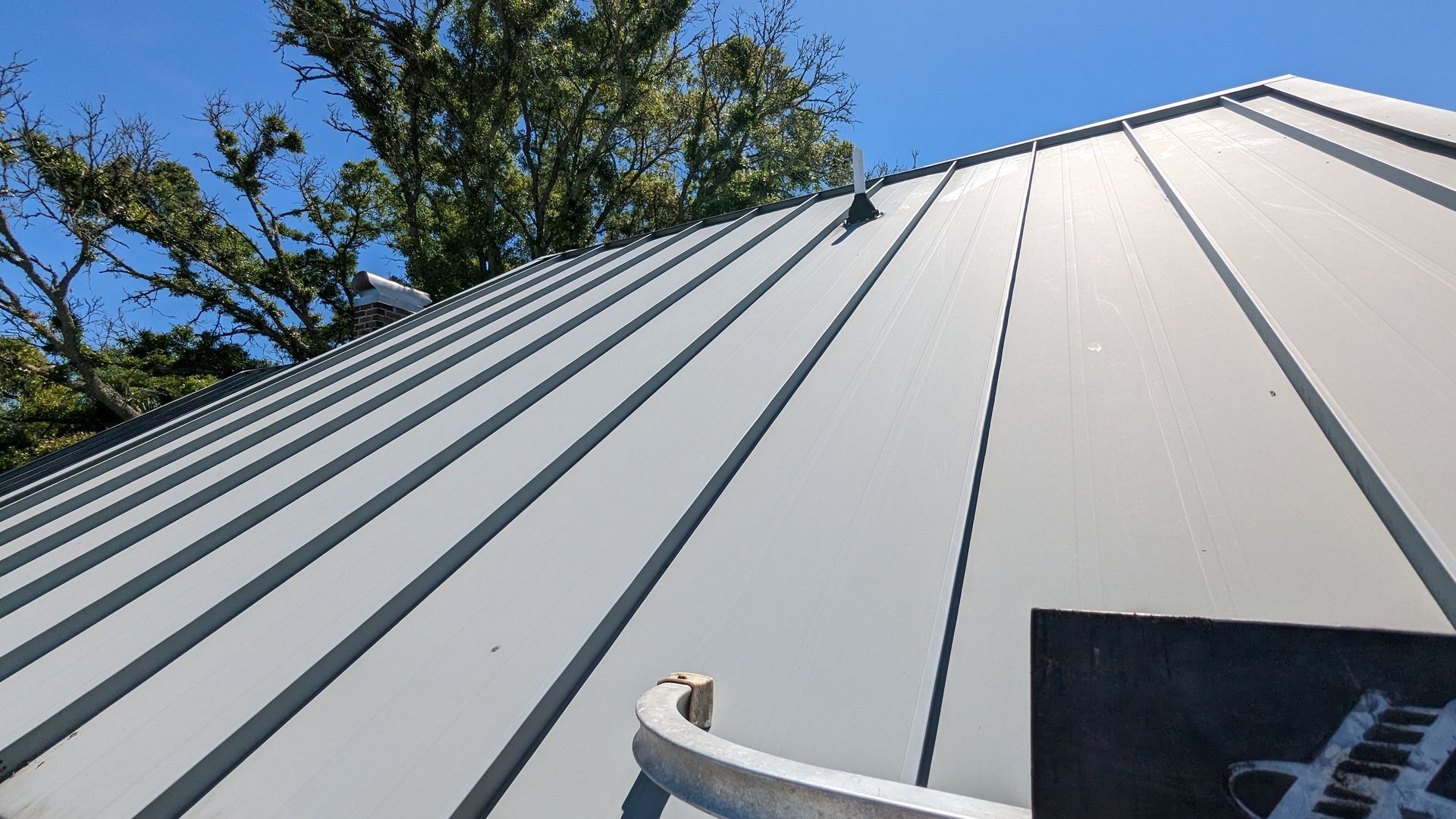 A light gray standing-seam metal roof viewed from a low angle against a clear blue sky, with trees visible at the top.