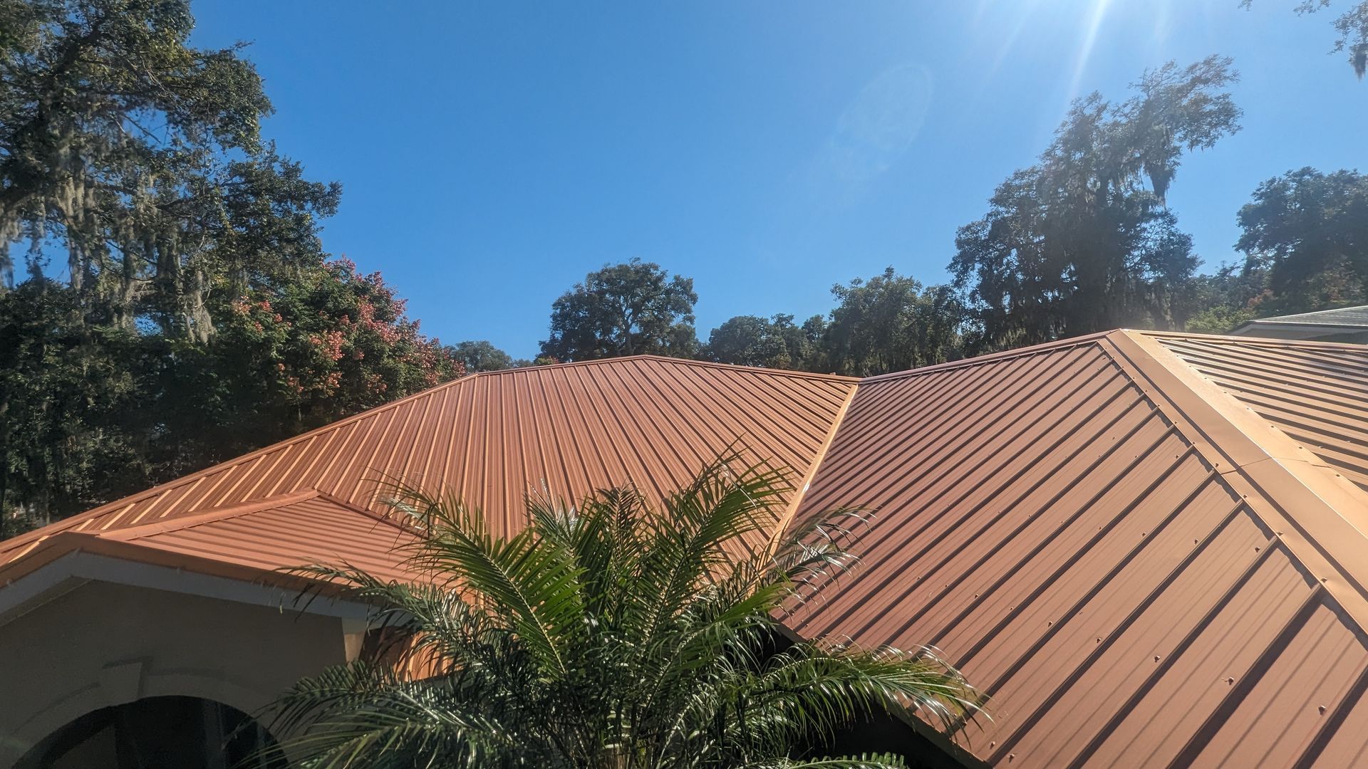 A sunlit view of a house roof featuring textured, reddish-brown metal shingles with a palm plant in the foreground.