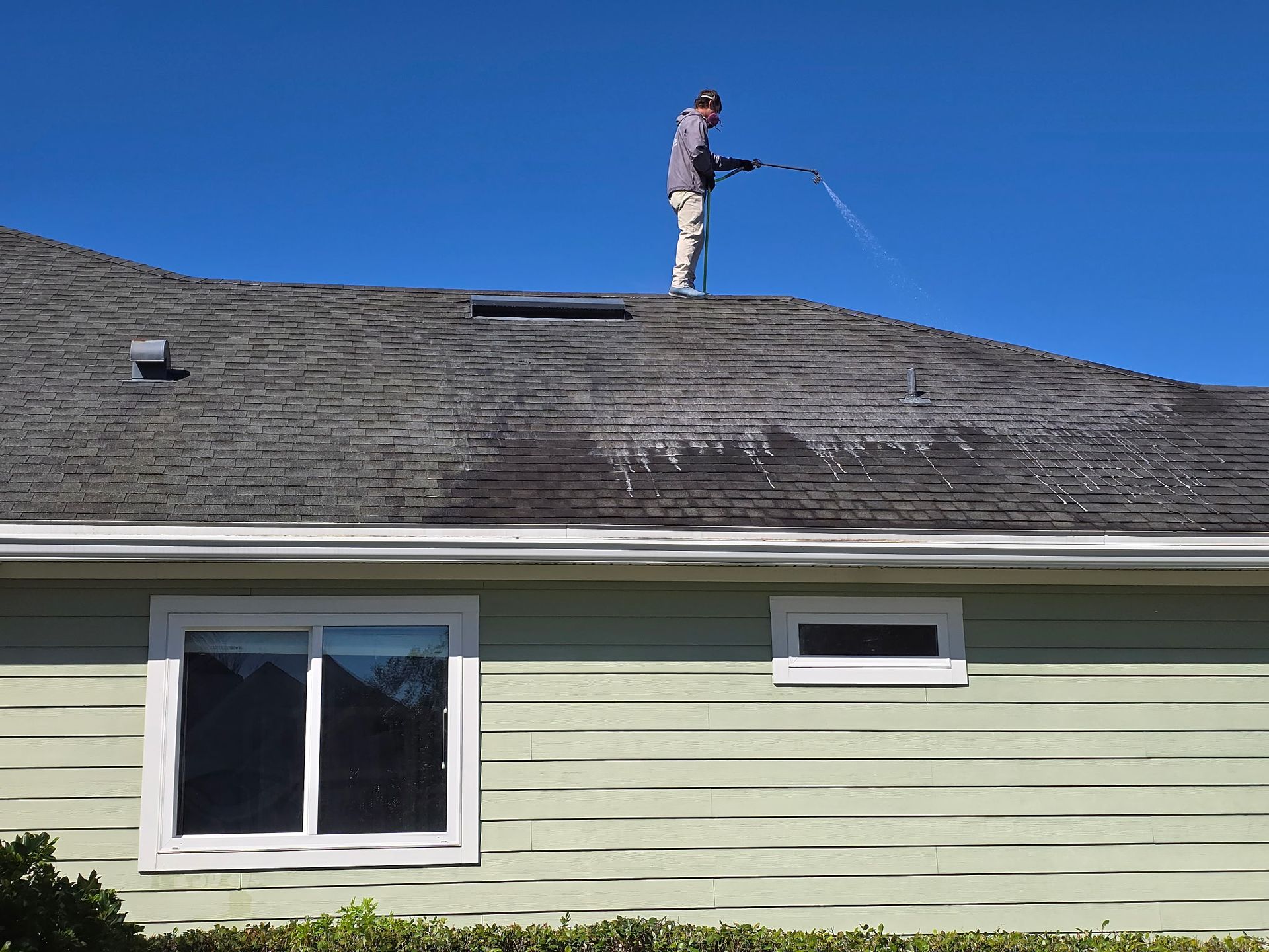 A person standing on a sloped roof under a clear blue sky, using a pressure washer to clean the shingles.
