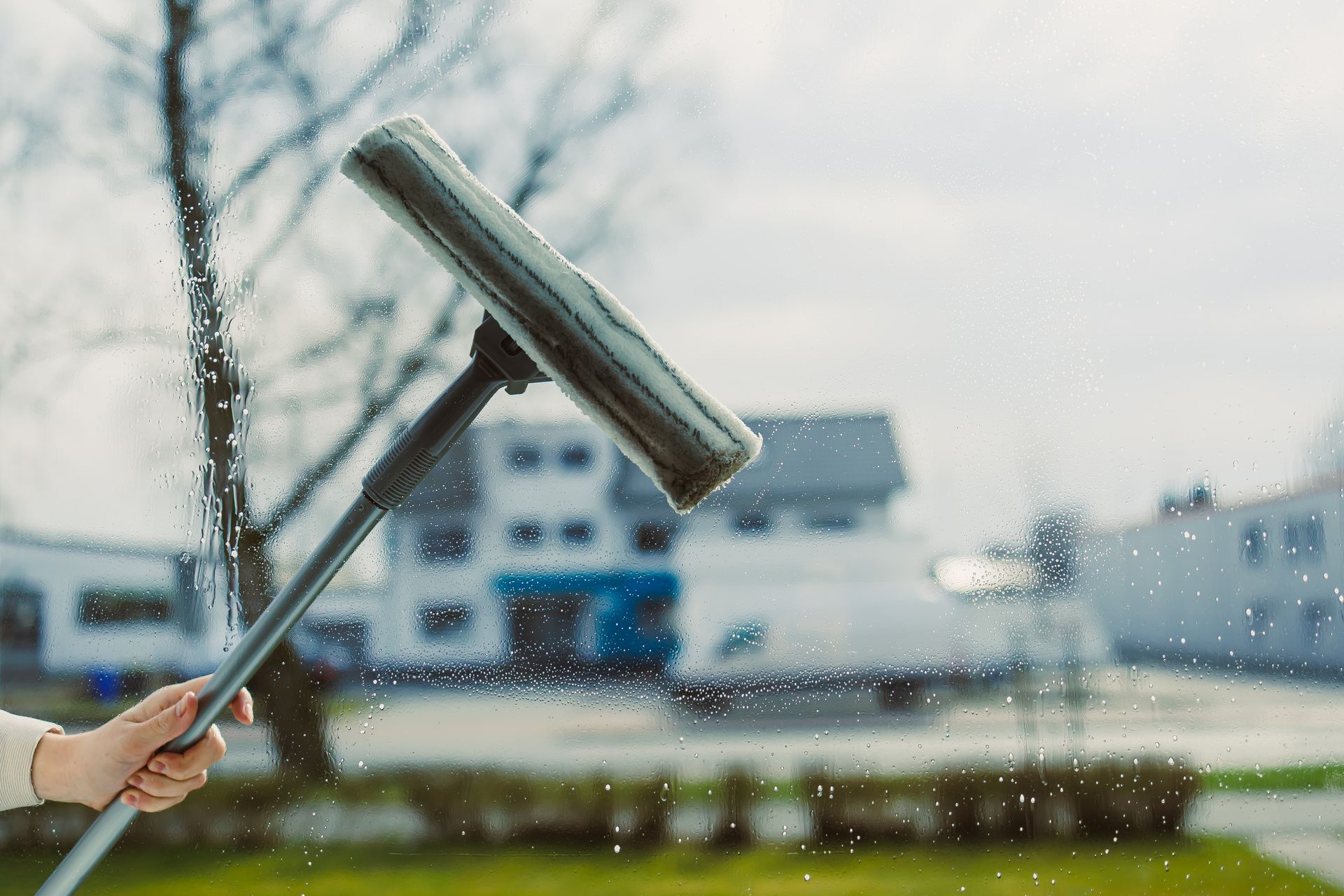 A hand holds a squeegee against a wet window, with a blurred building and green lawn visible in the background.
