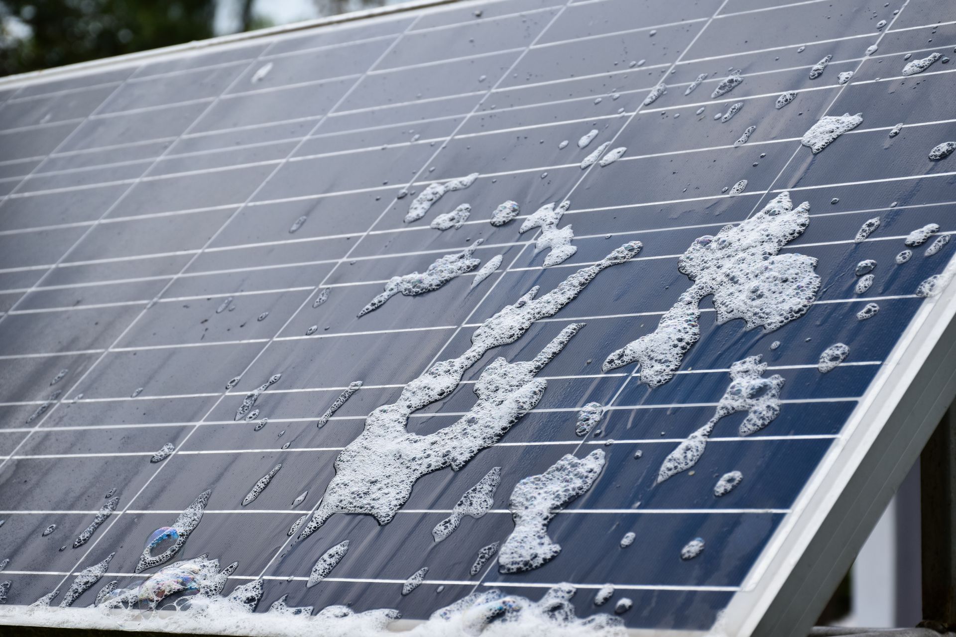A close-up of a solar panel being cleaned with foamy, white soap suds on its blue, gridded surface.