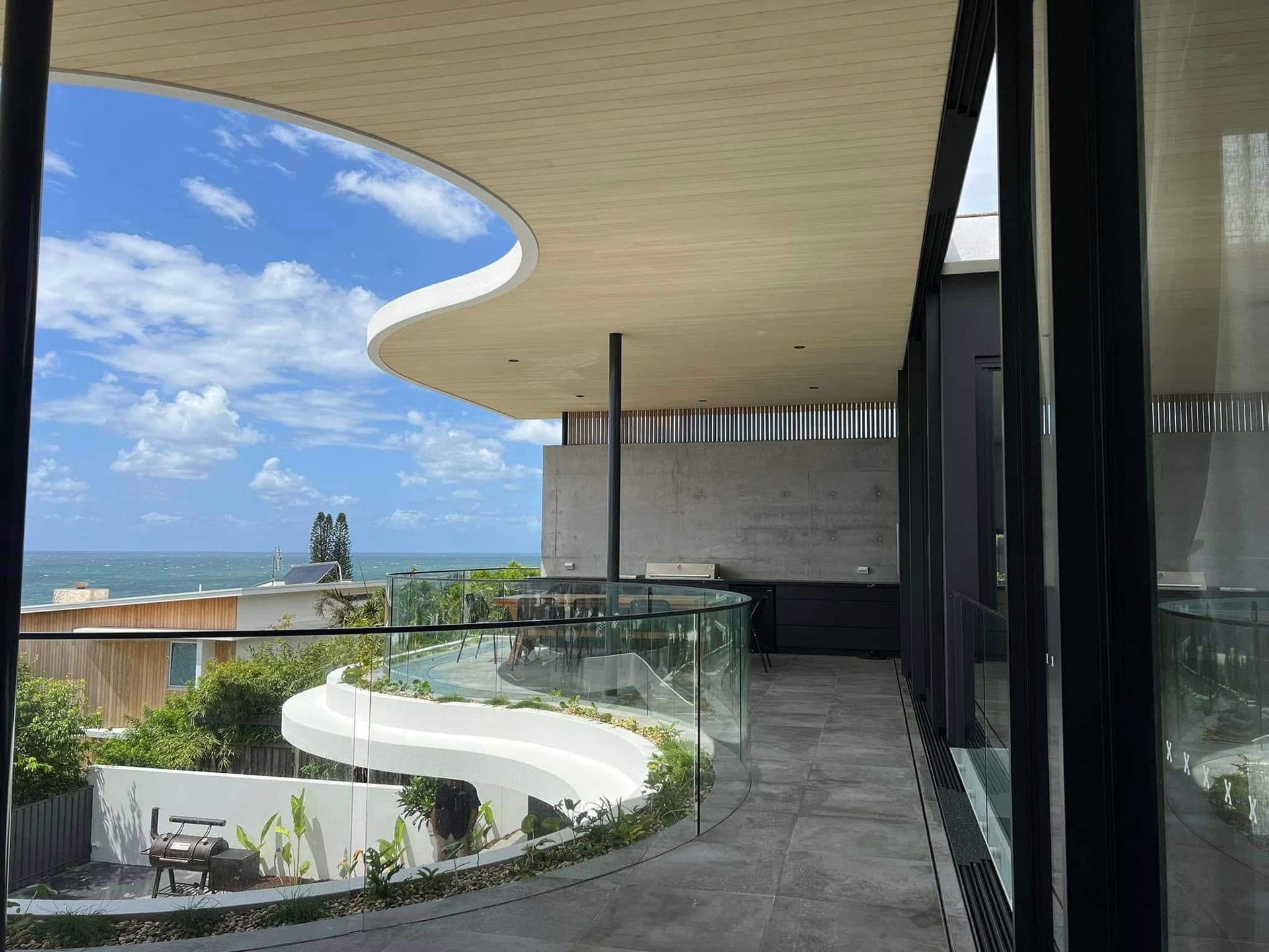Balcony with ocean view, curved roof, glass railing, and blue sky. — Frameless Shower Installations in Caloundra, QLD