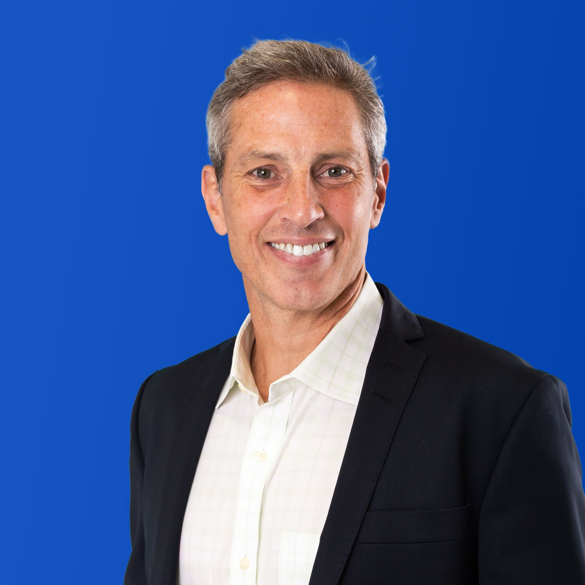 A man in a suit and plaid shirt is smiling in front of a yellow circle.