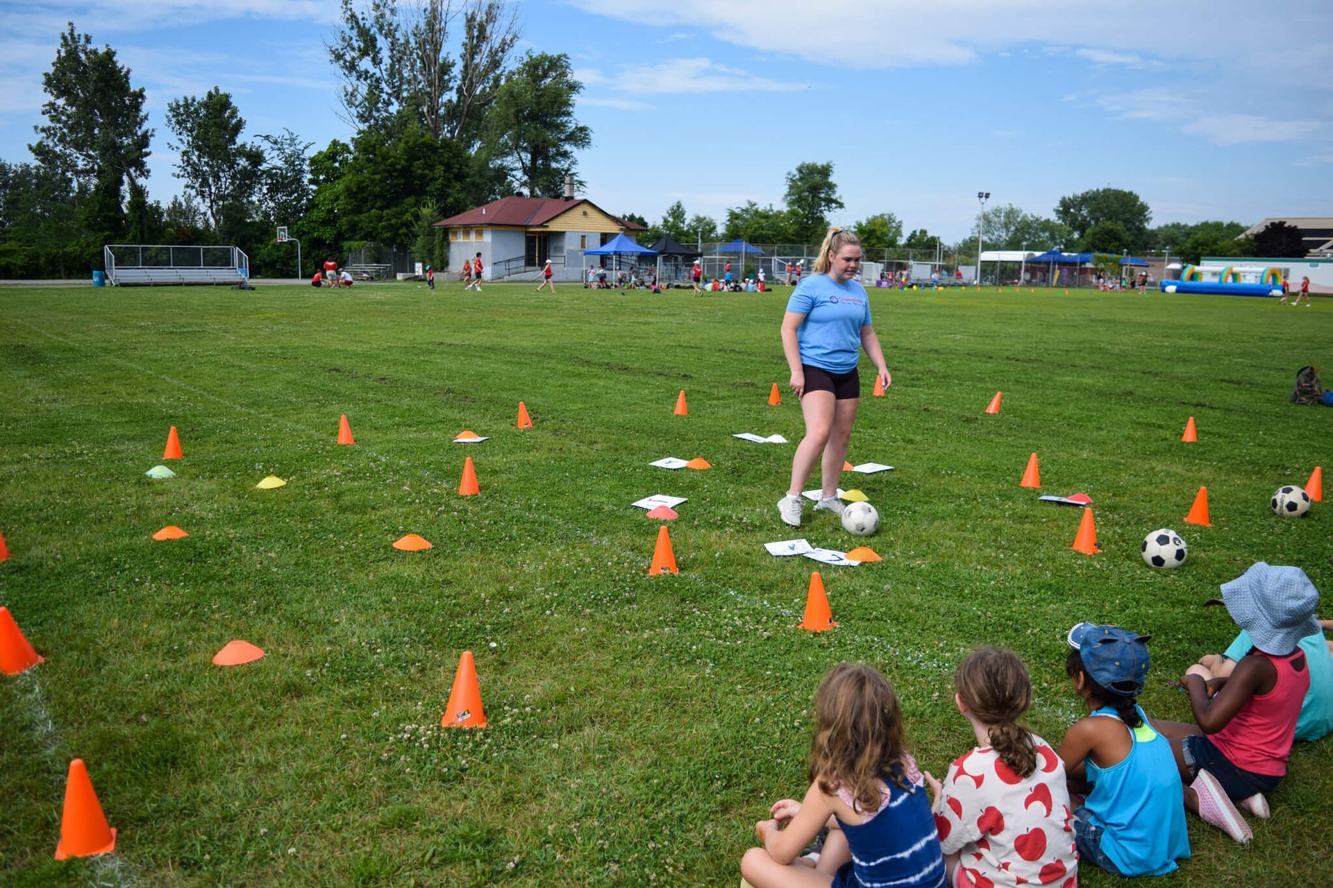 A group of children are sitting on the grass watching a woman play soccer.