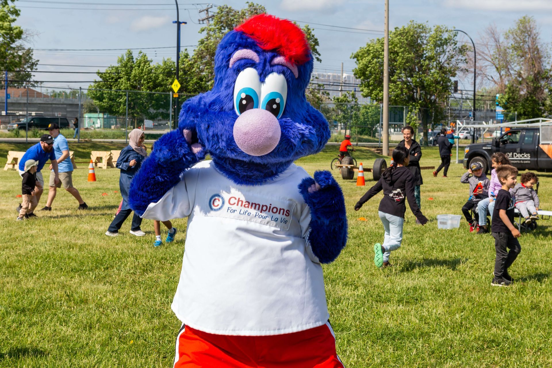 A blue mascot is standing in a field with children playing in the background.