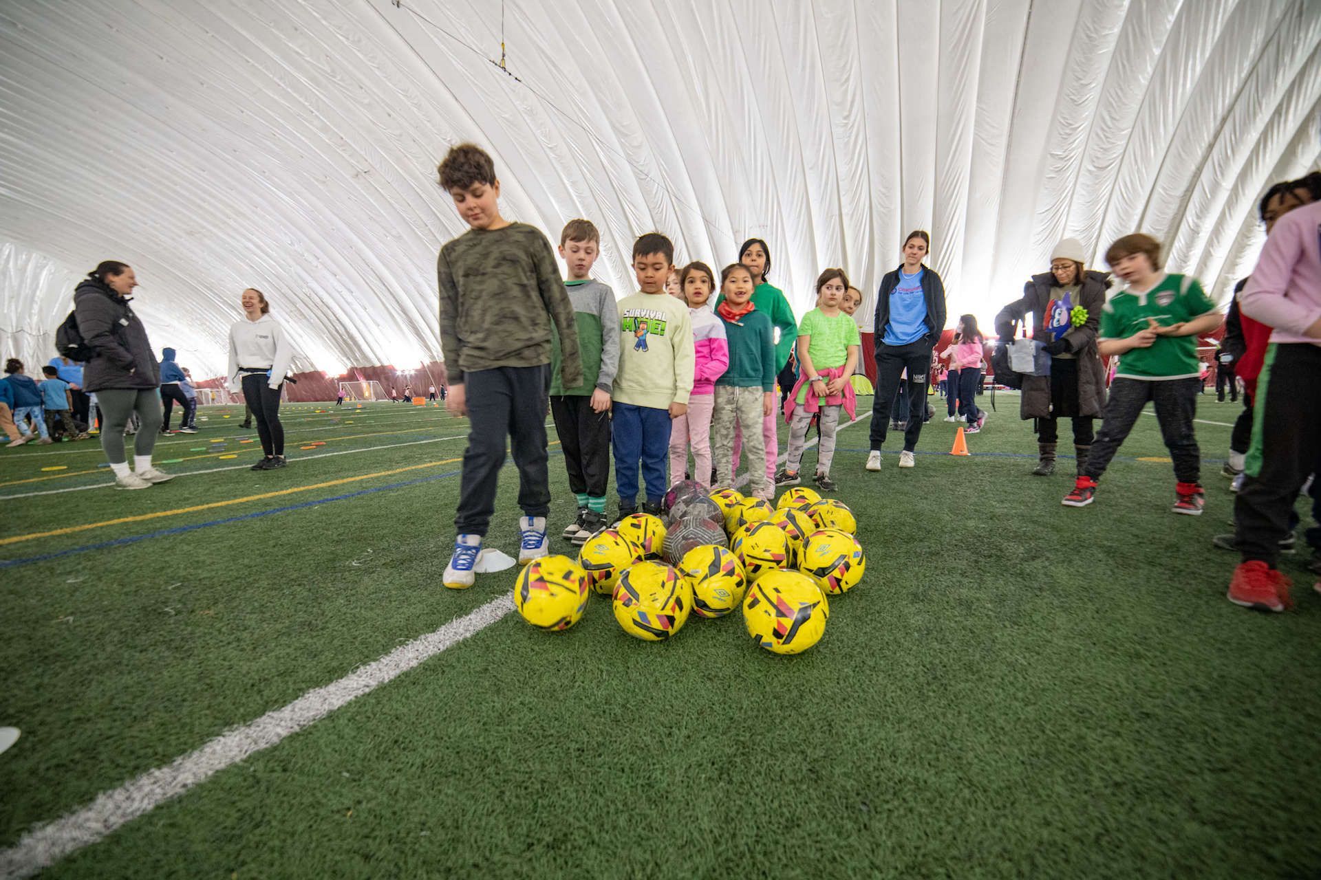 A group of children are standing next to a pile of soccer balls on a field.