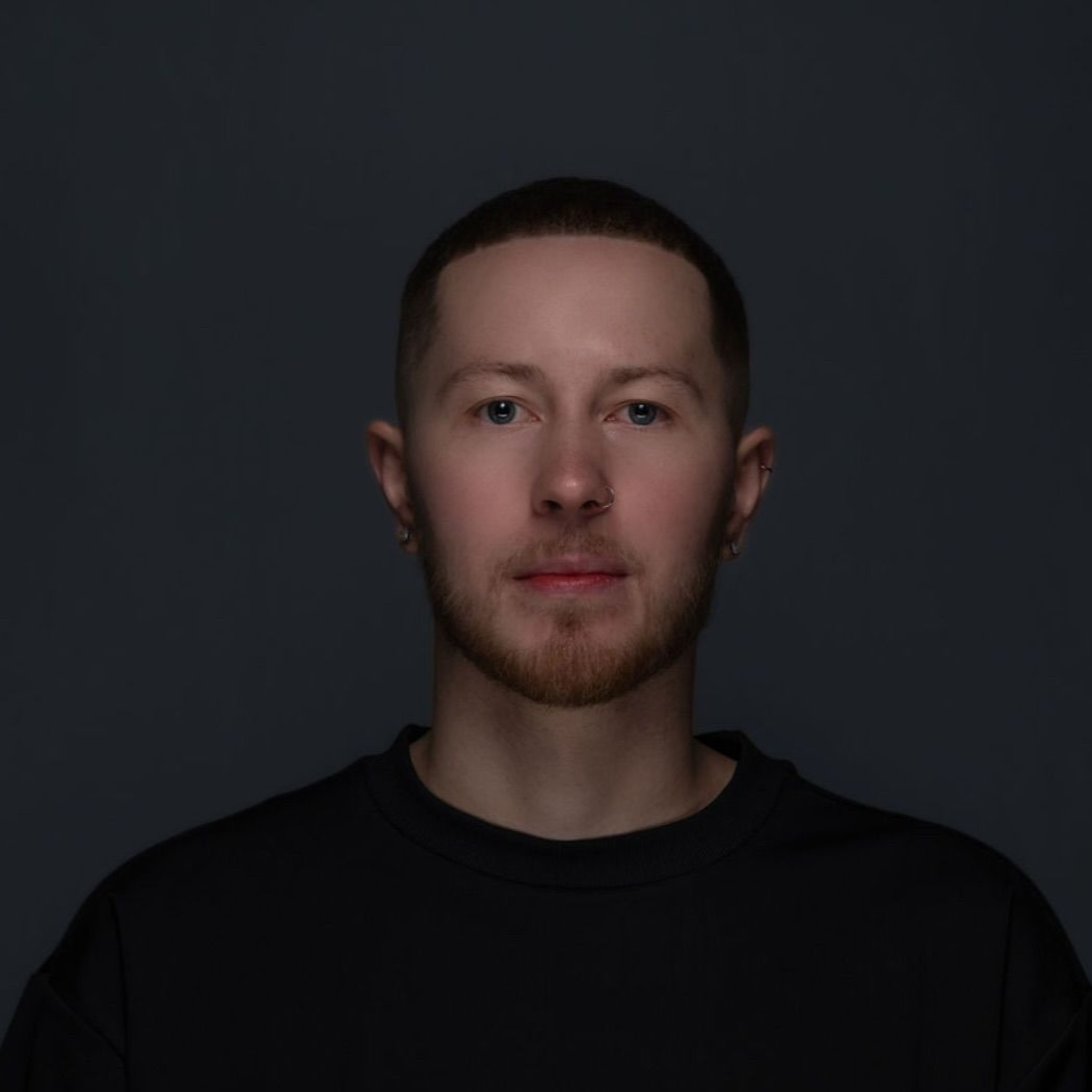 Man with short hair and stubble in a dark shirt, lit against a dark background.