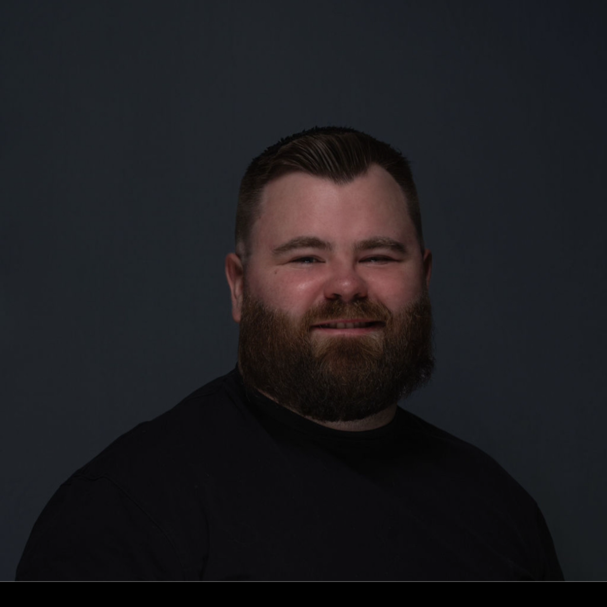 Man with a full beard smiles, wearing a black shirt against a dark background.