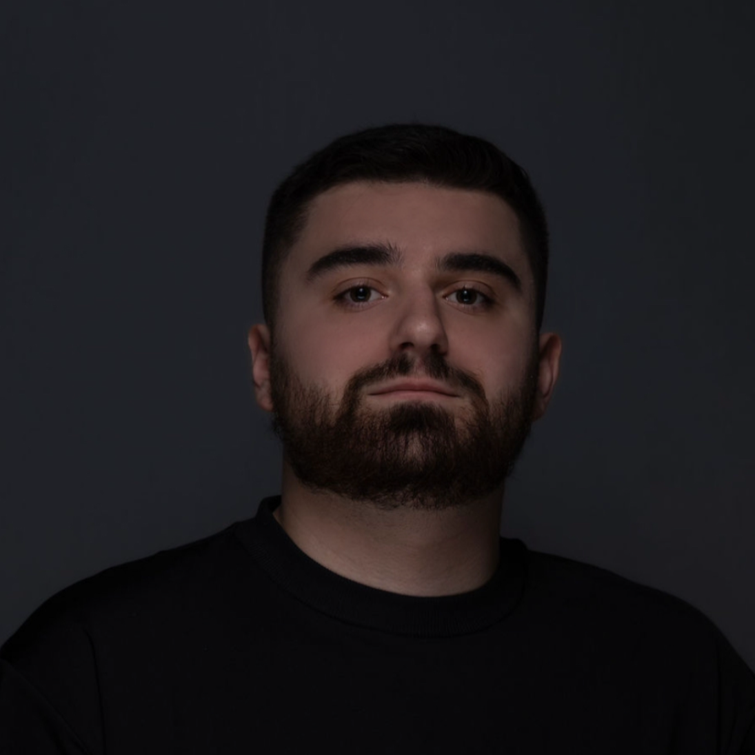 Man with dark hair and beard against a gray background, looking at the camera.