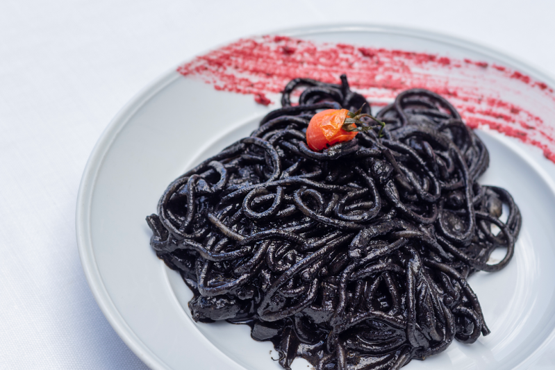 Spaghetti neri su un piatto bianco, guarnito con un singolo pomodorino rosso e una spalmata di salsa rossa.