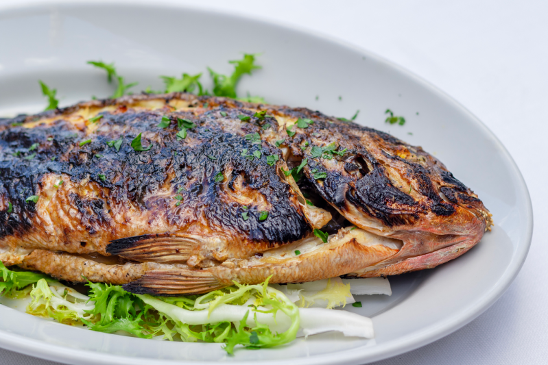 Pesce intero grigliato su un piatto bianco con verdure, qualche erba aromatica e uno sfondo bianco.