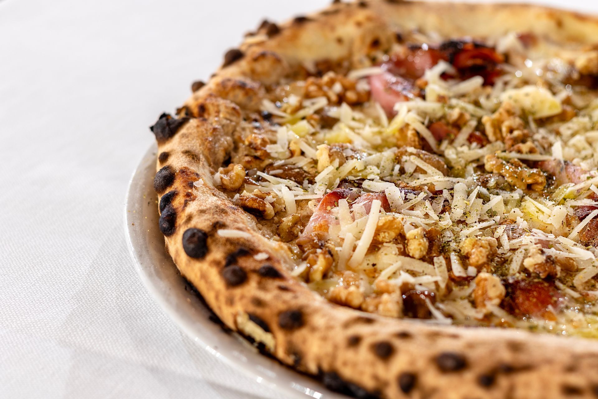 Primo piano di una pizza su un piatto bianco, con crosta bruciacchiata e condimenti tra cui noci e formaggio.
