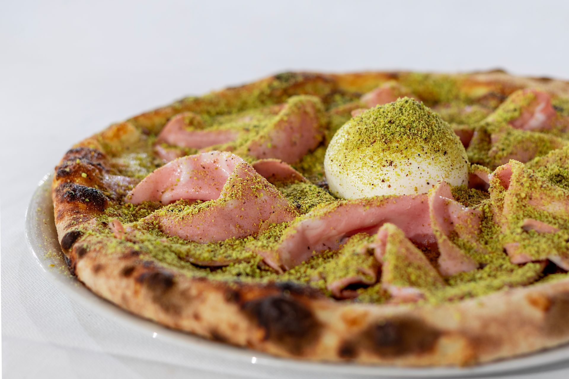 Pizza con pistacchi, prosciutto e una pallina di burrata su un piatto bianco.