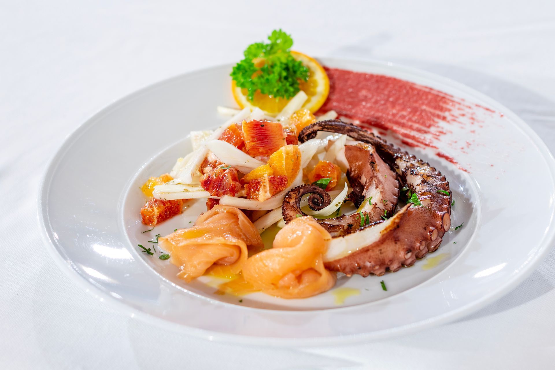 Piatto di polpo grigliato, arance, finocchio e salsa rossa, guarnito con prezzemolo e una fetta d'arancia.