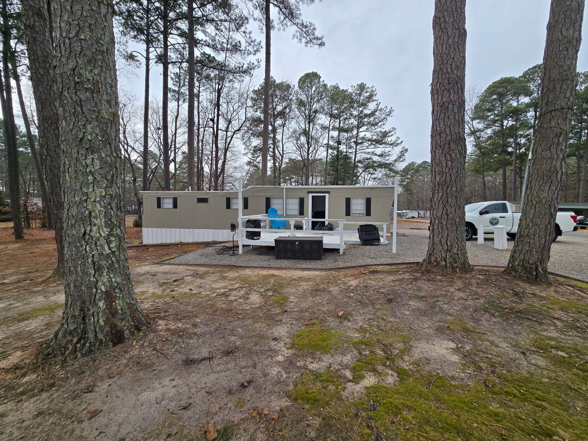 Mobile home with a small deck and two trees in the foreground. Gray and white colors. Overcast day.
