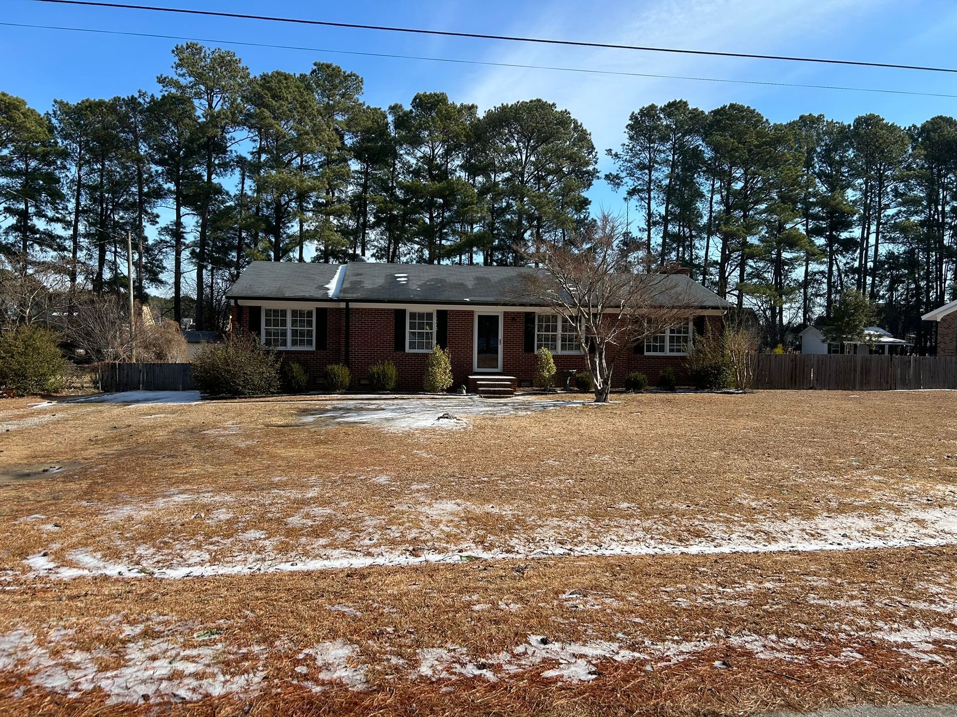 Brick ranch house with brown lawn, trees in the background, under a blue sky.