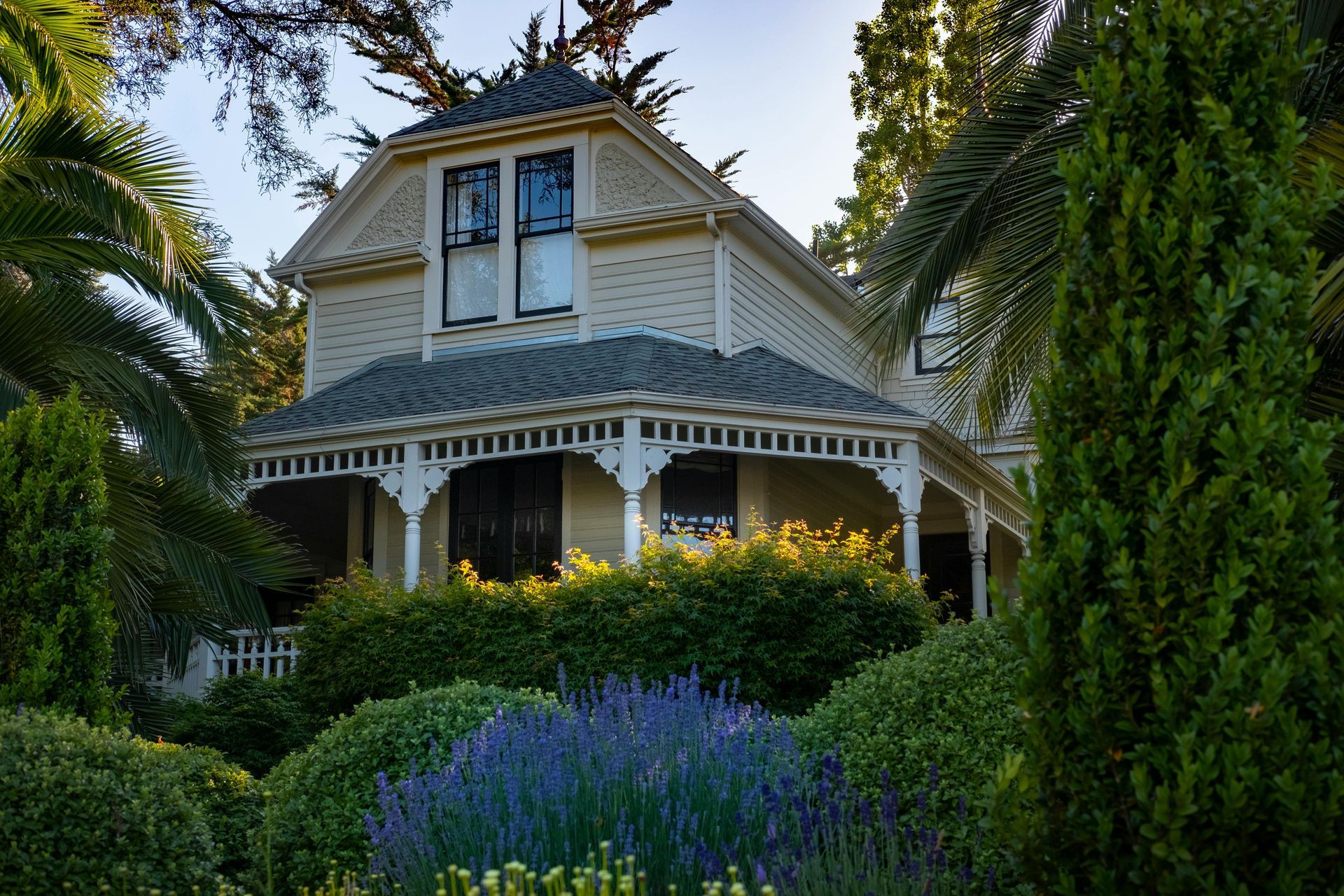 Victorian-style house with light-colored siding, surrounded by lush green and purple landscaping.