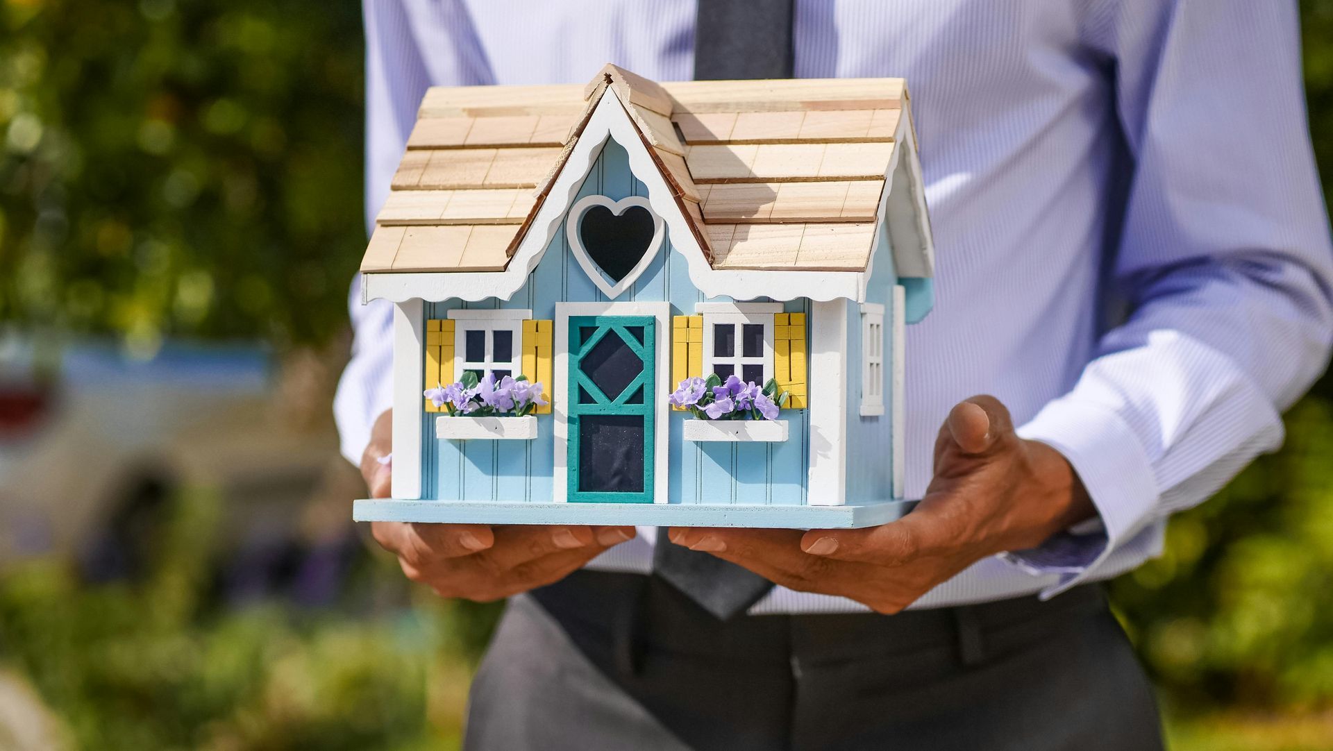 Person holding a miniature blue house with yellow shutters; blurred outdoor background.