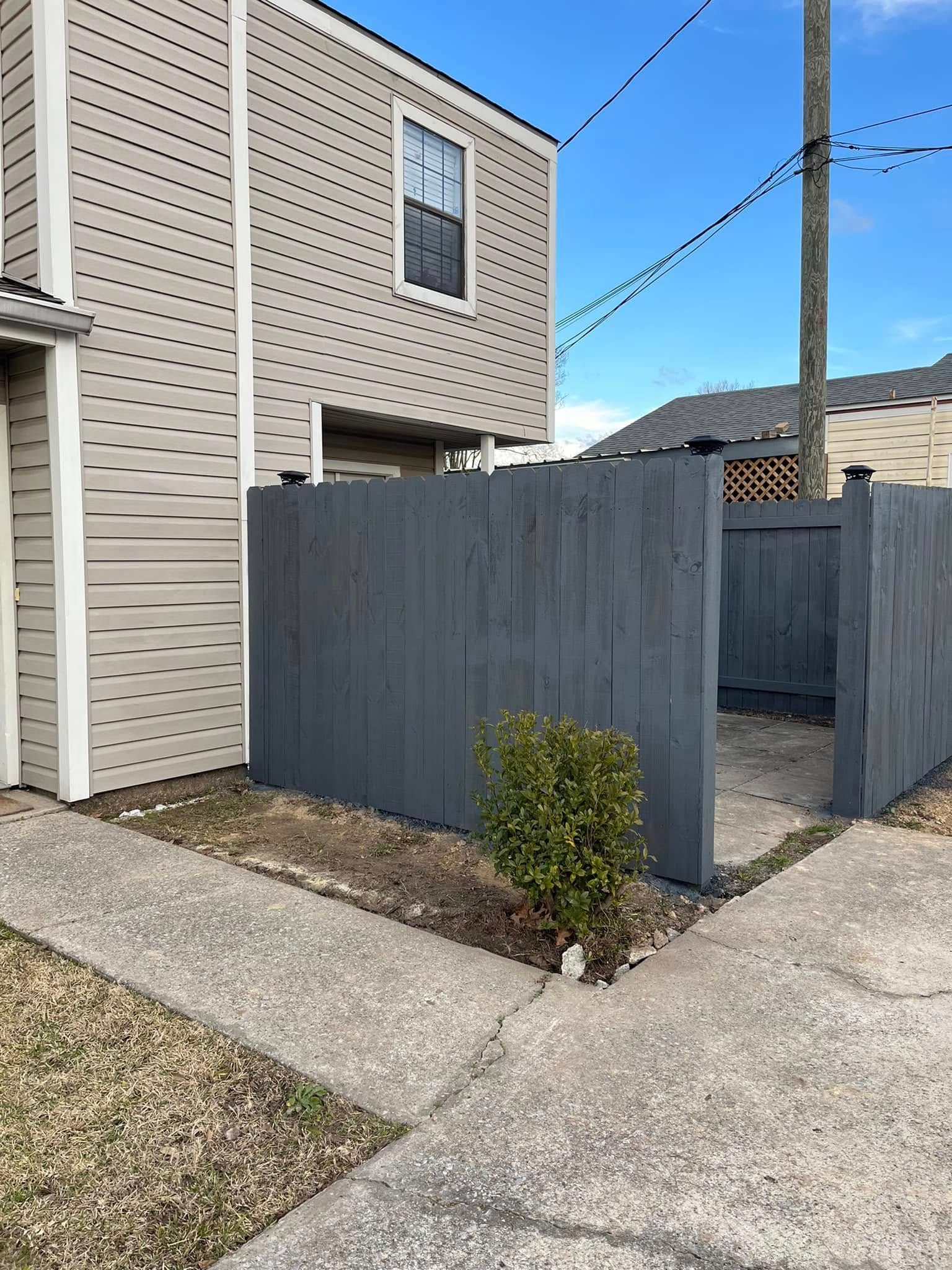 Exterior view of a townhome with a gray fence and small green bush on a concrete path under a blue sky.