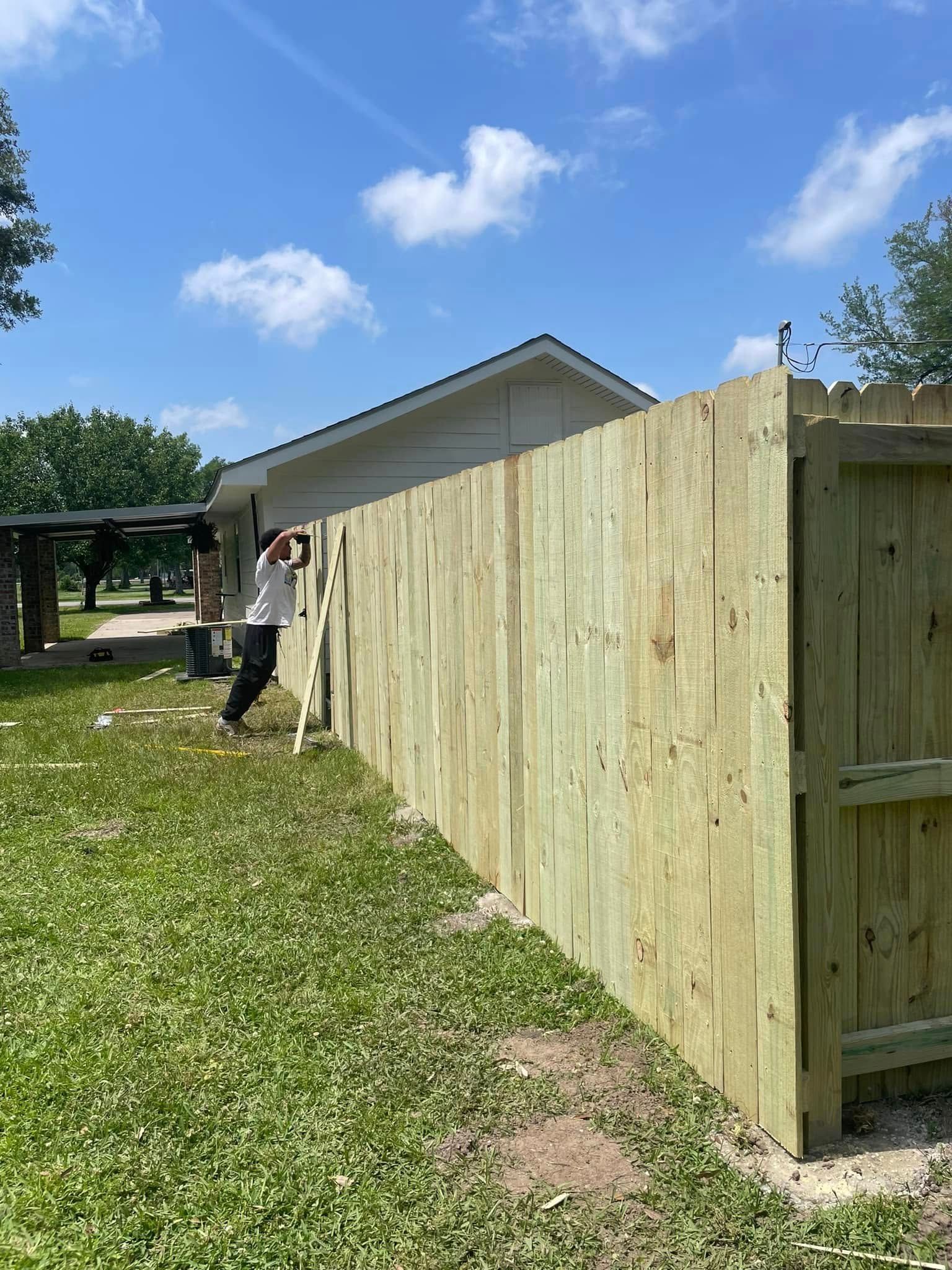 Man building a wooden fence outdoors on a sunny day. Green grass, blue sky, and a white building in the background.