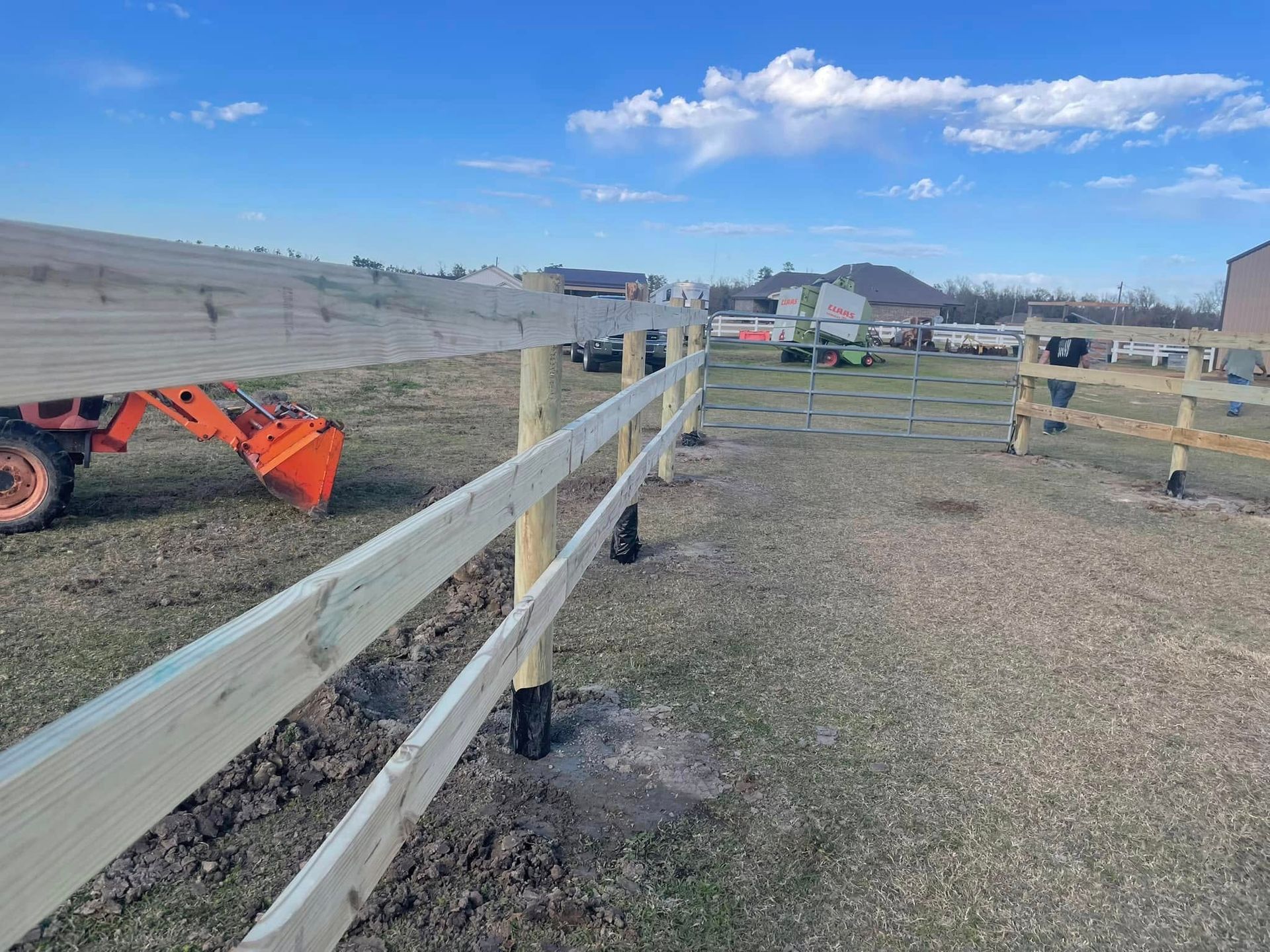A wooden fence being built in a grassy field on a sunny day. An orange tractor sits nearby.