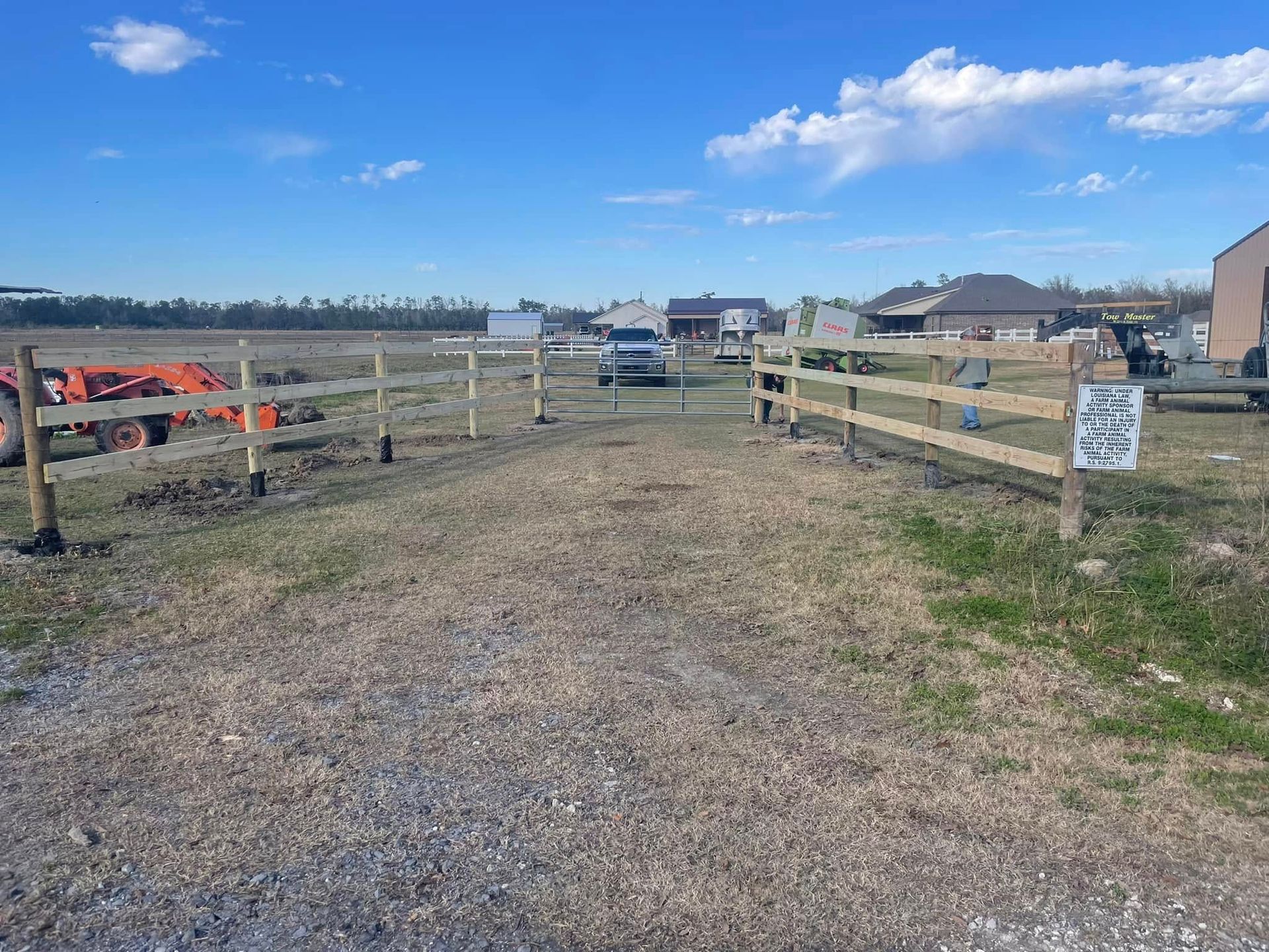 A gravel driveway leads to a wooden fence entrance in a field under a blue sky.