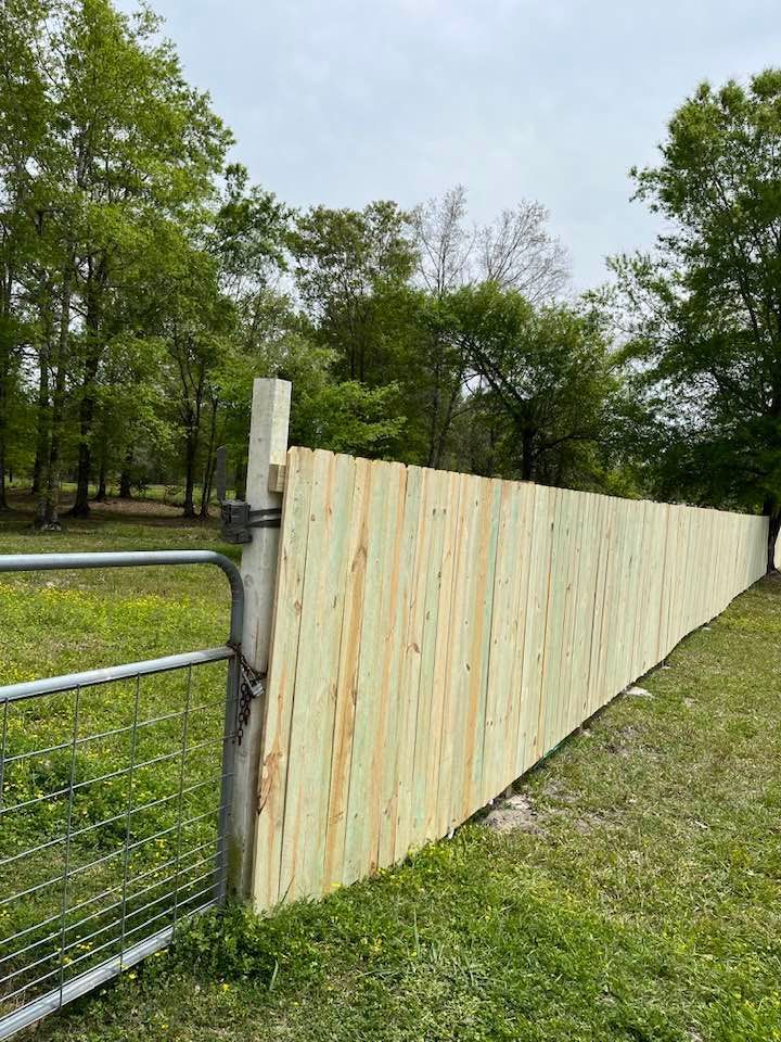Newly constructed wooden fence with gate in a grassy field, trees in background.