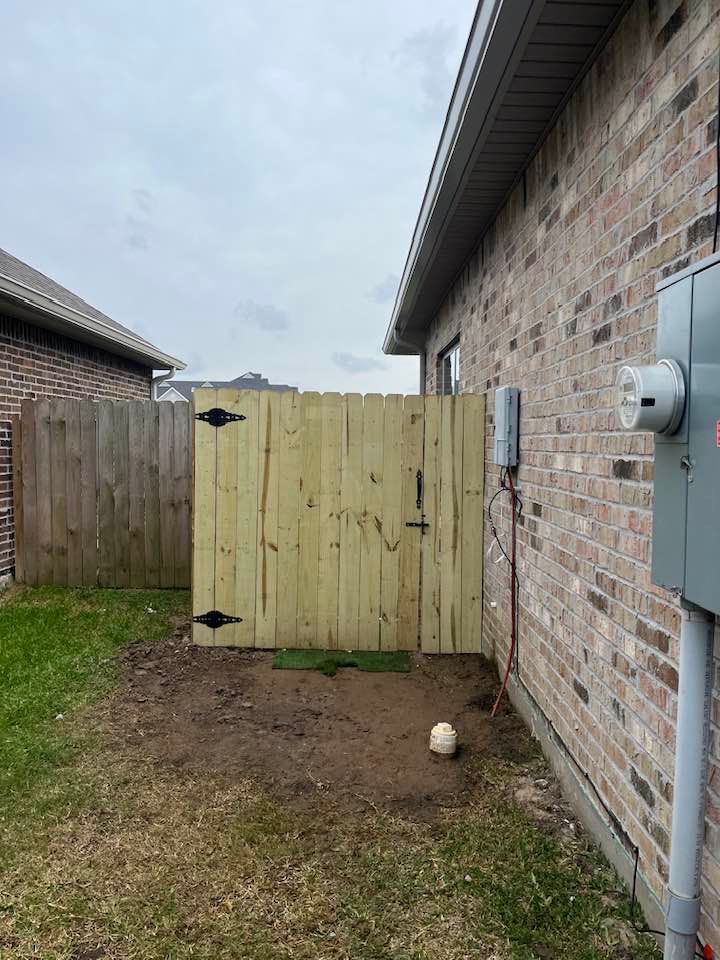 Wooden fence gate between two brick buildings. Grass and dirt ground. Electrical box on the right.