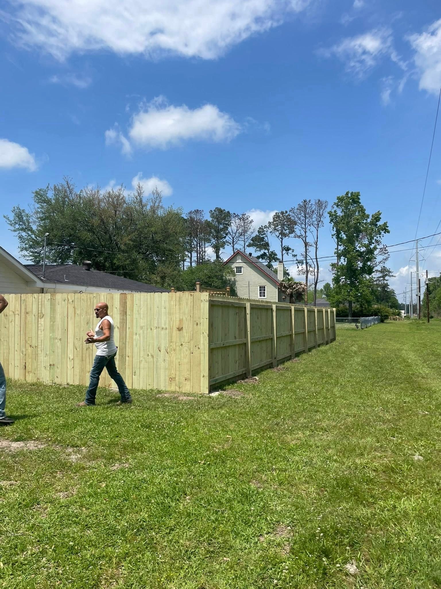 Wooden fence being built in a grassy yard, man walking. Cloudy sky.