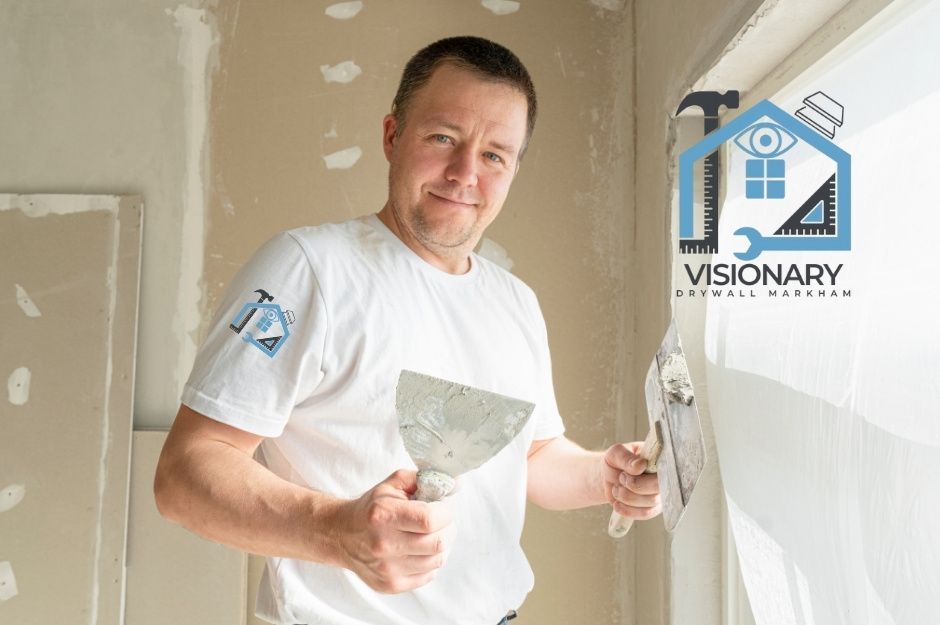 A drywall repair company worker, wearing a white t-shirt, holds two putty knives while standing in front of unfinished drywall with joint compound patches. Middle-aged man with short brown hair smiles during commercial construction renovation work near Concession Rd 6, and Westney Rd, Pickering, ON.
