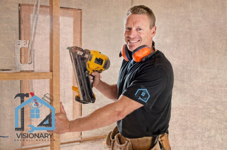 A drywall repair handyman smiling while using a framing nailer on wooden studs at a residential construction site, wearing orange hearing protection and a tool belt near Wildwood Ave, and Newman Ave, Richmond Hill, ON.