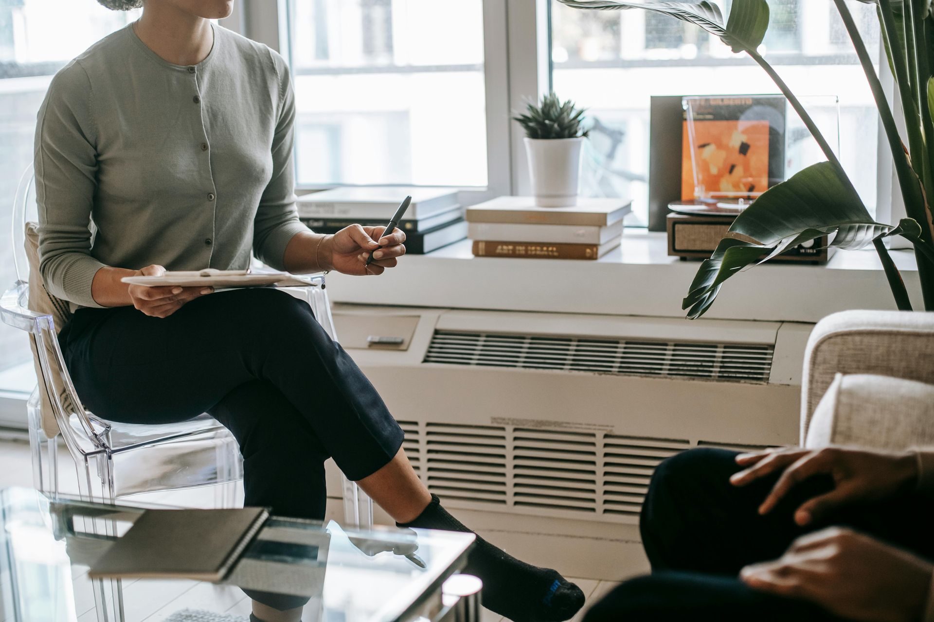 One person sitting in a chair, holding a notepad in one hand, speaking with another person sitting 