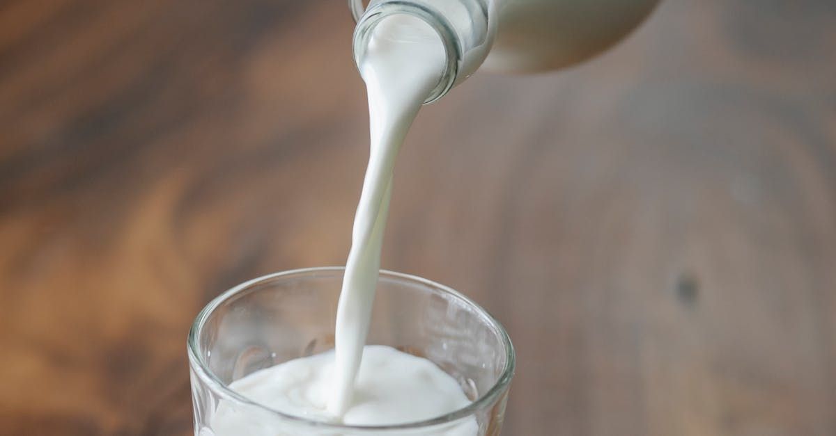 A bottle of milk is pouring milk into a glass that is sitting on a solid surface like a table. 