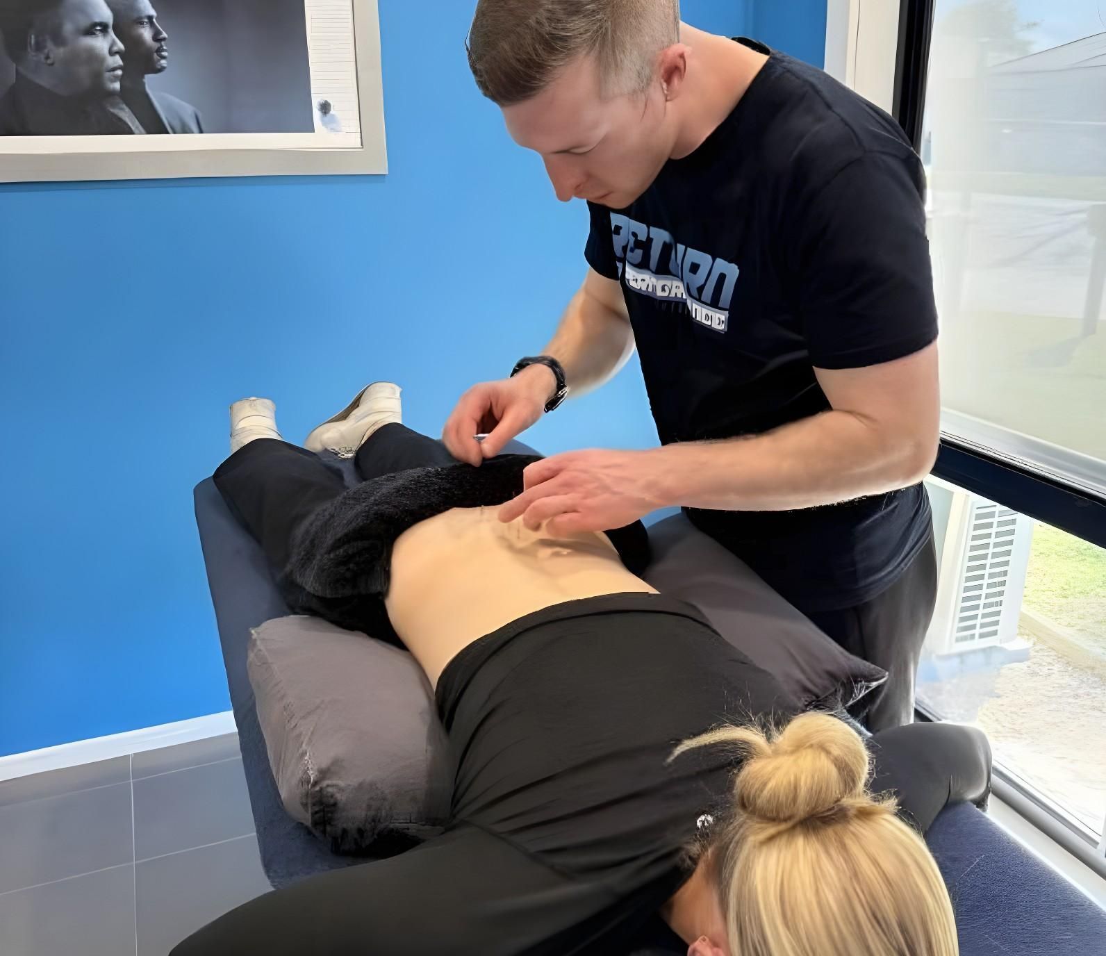 A Therapist Examining a Patient's Abdomen on a Medical Table. Blue Wall, Black Clothing — Return to Performance Physiotherapy in Epsom, VIC