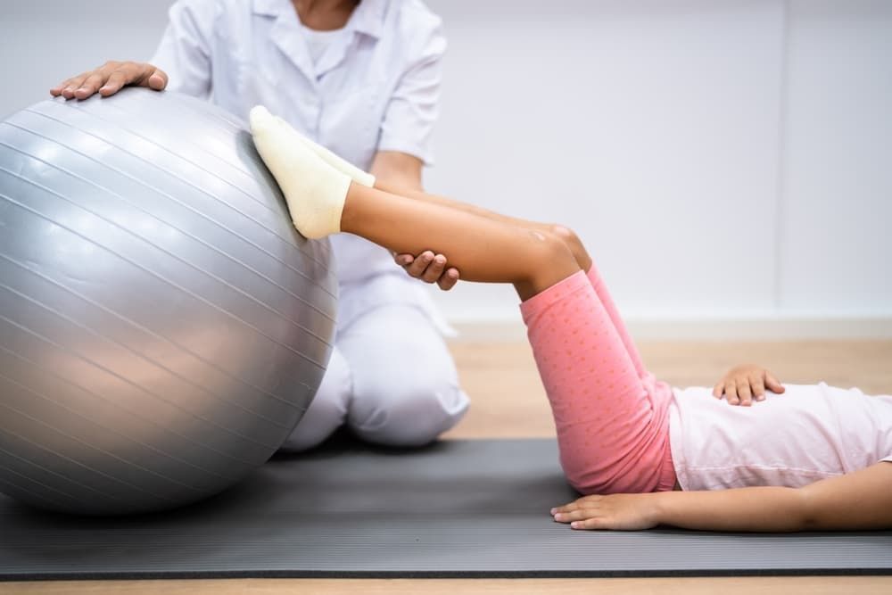 A Young Girl is Laying on a Yoga Mat While a Nurse Holds an Exercise Ball — Return to Performance Physiotherapy in Epsom, VIC