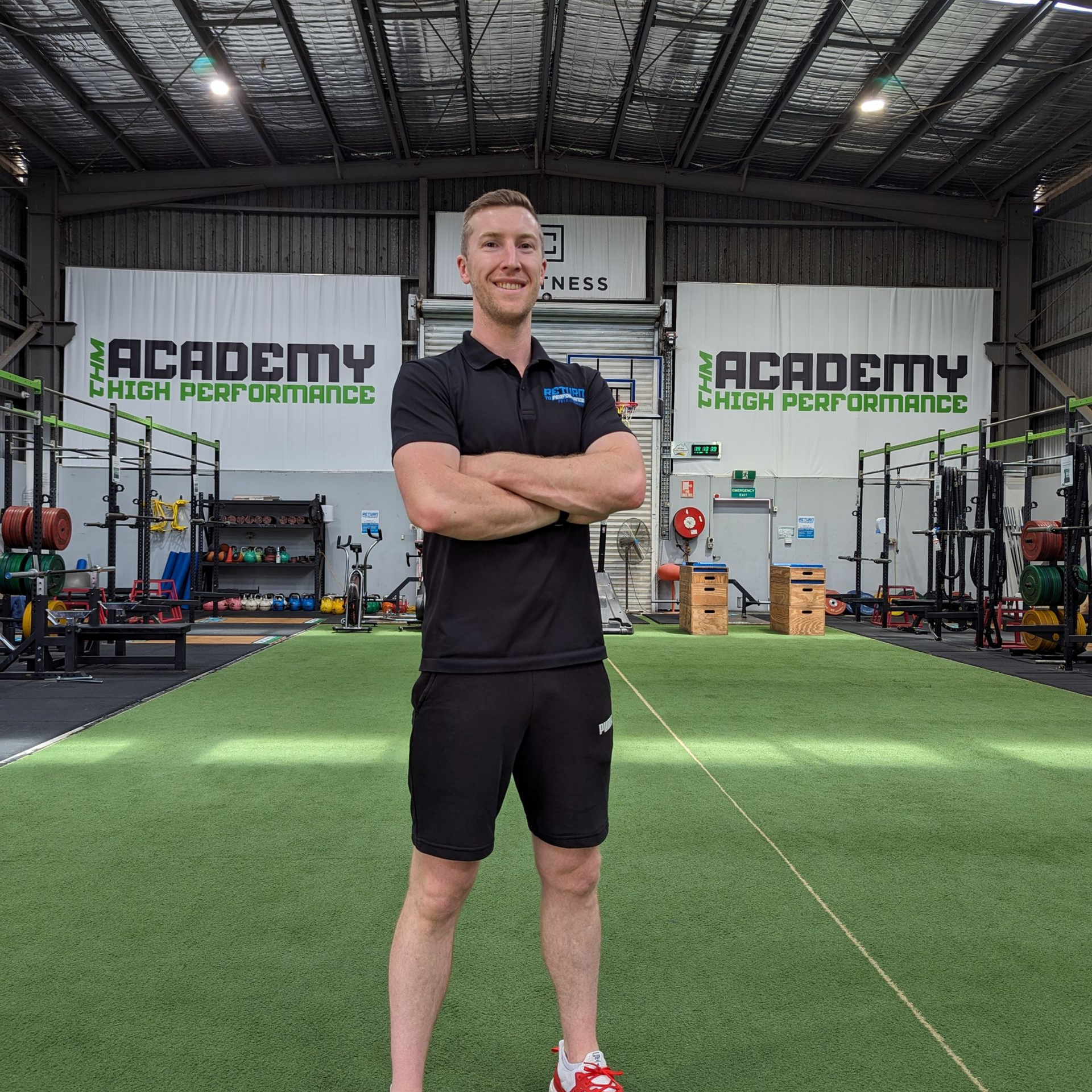 Man With Arms Crossed in a Gym, Wearing Black Shorts and Polo — Return to Performance Physiotherapy in Epsom, VIC