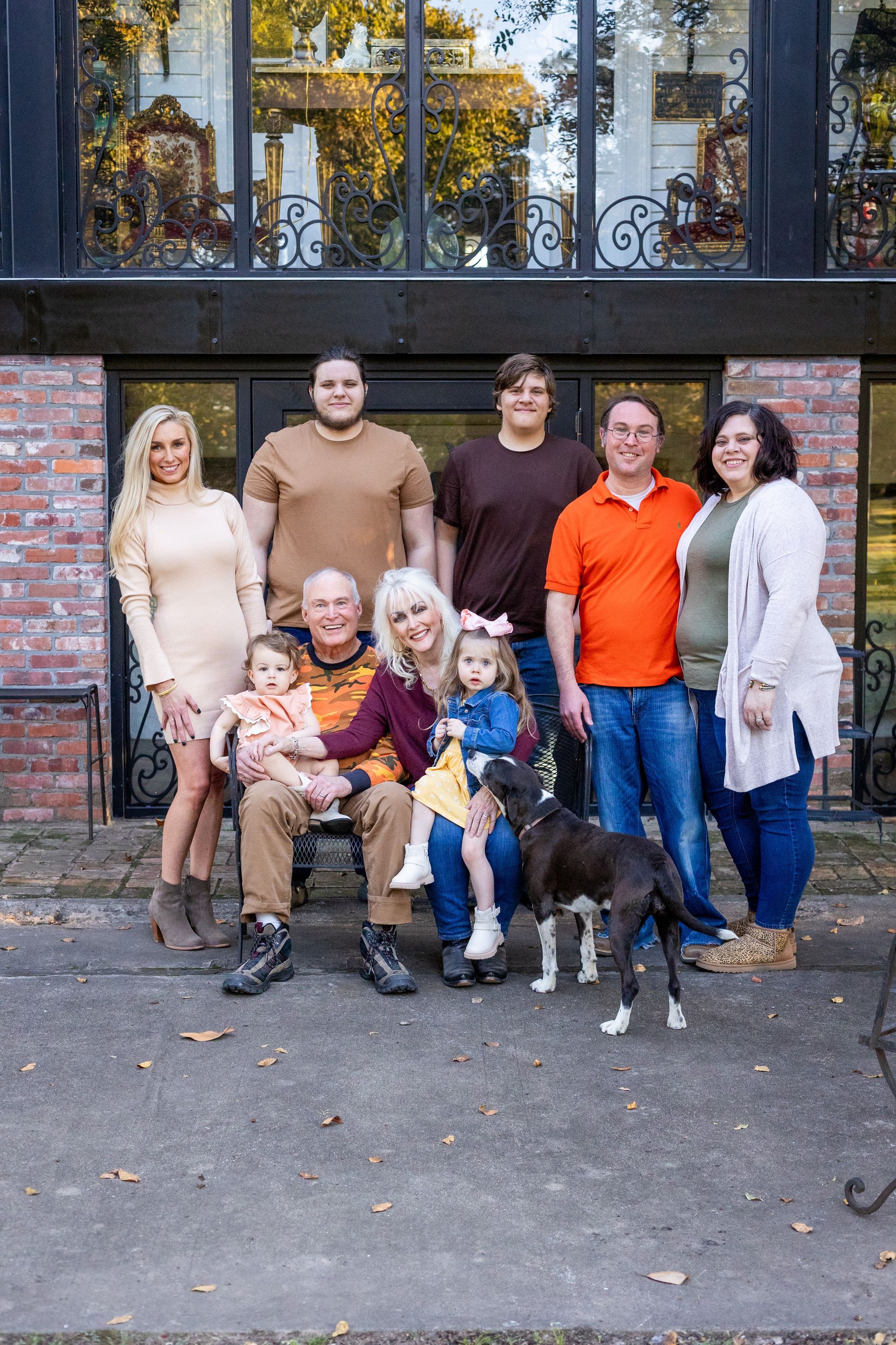 Group of seven people posing outdoors against a brick wall, with two small dogs in front.