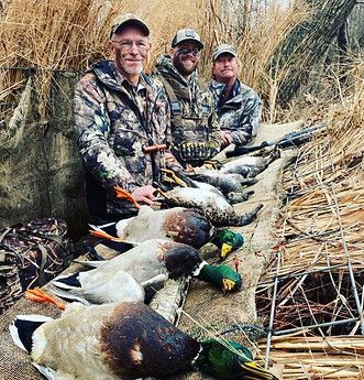 Three hunters in camouflage pose with several ducks laid out in a marsh.
