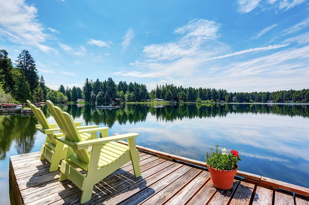 Green Adirondack chairs on a wooden dock by a calm lake with a potted plant and blue sky reflections.