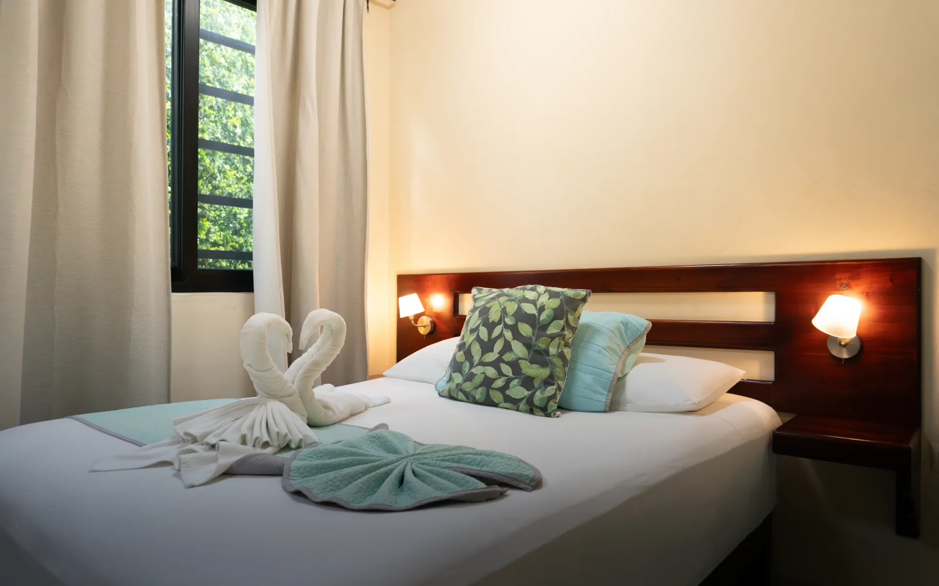 Cama de hotel impecablemente hecha con cisnes de toalla, almohadas verdes y lámparas.