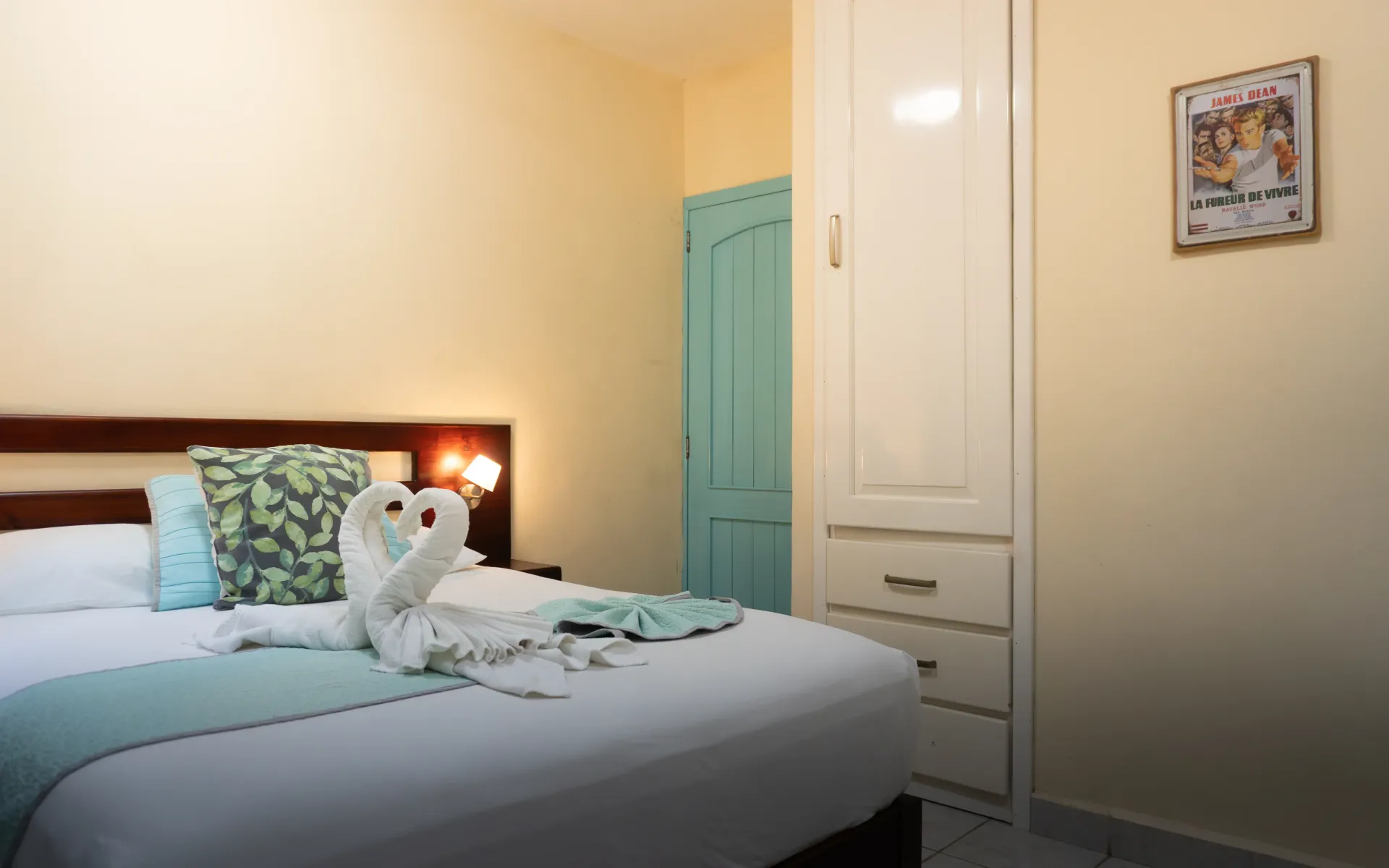 Cama bien hecha con cisnes de toalla blancos y ropa de cama color turquesa en un dormitorio color crema con armario.