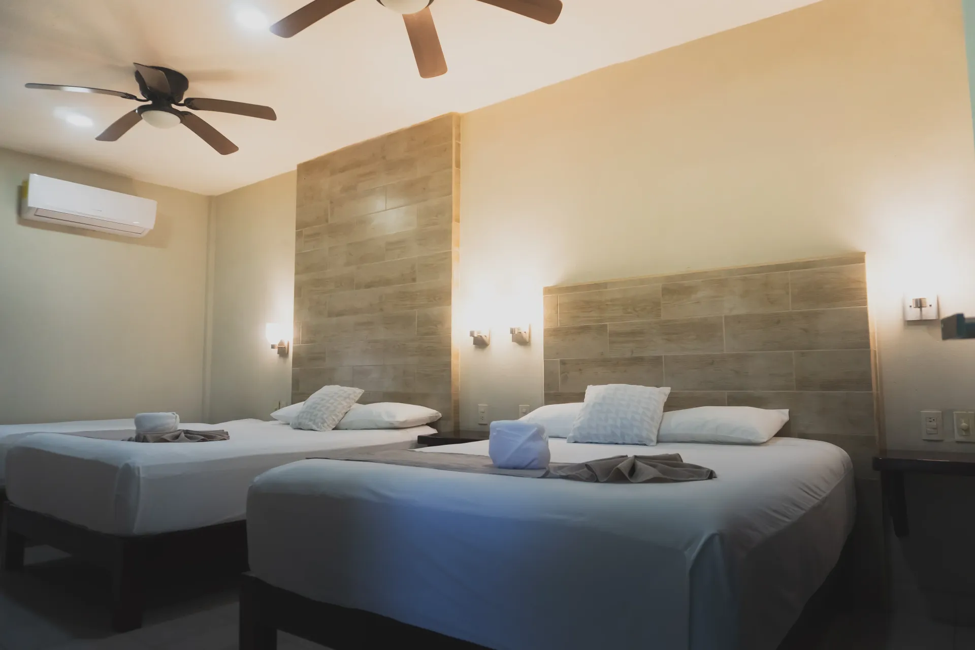Habitación de hotel con dos camas bien hechas, apliques de pared, ventilador de techo y aire acondicionado de pared.