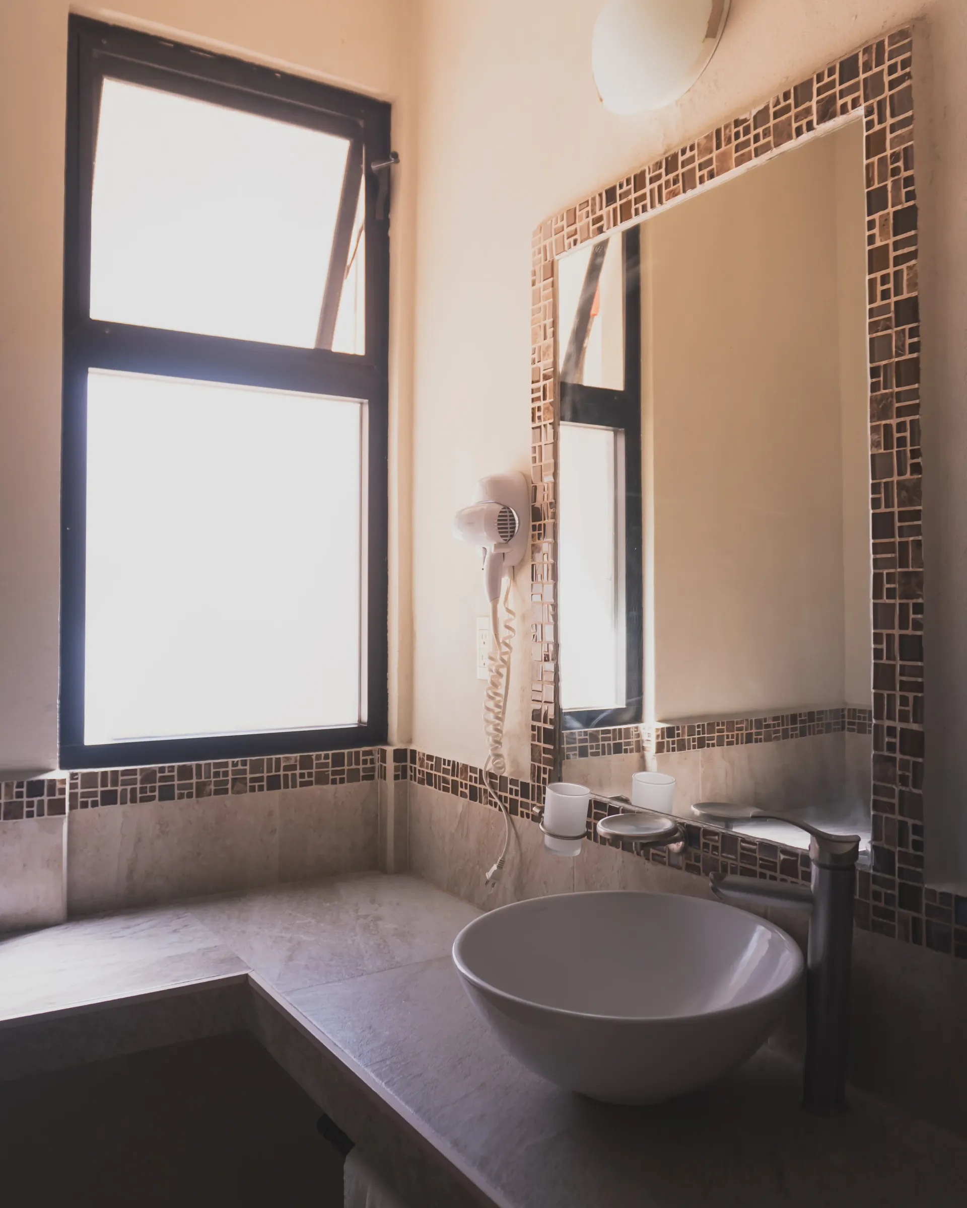 Baño moderno con lavabo redondo, espejo grande, paredes alicatadas y una ventana luminosa.