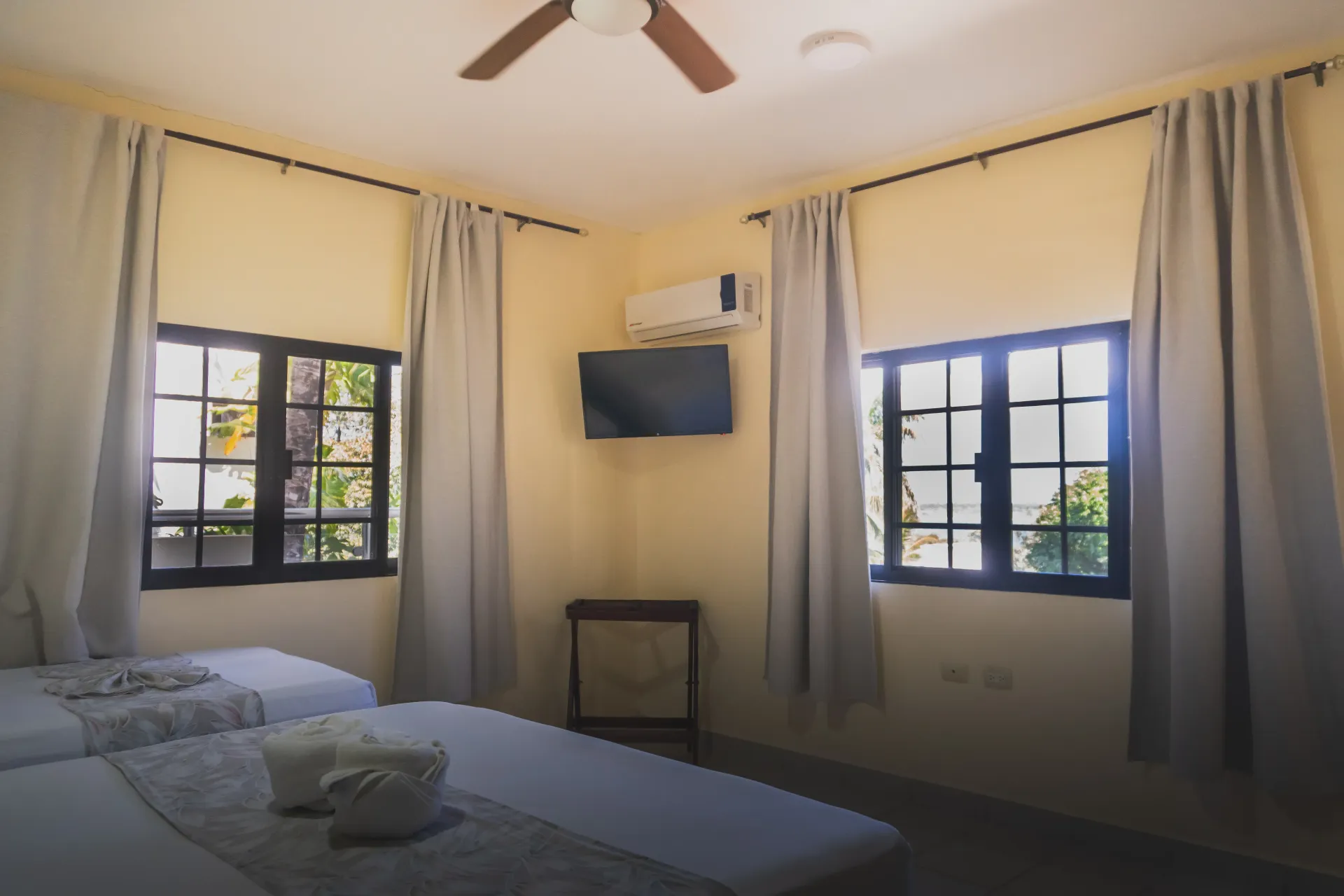 Habitación luminosa con dos camas individuales, dos ventanas, cortinas y un televisor montado en la pared sobre una mesita.